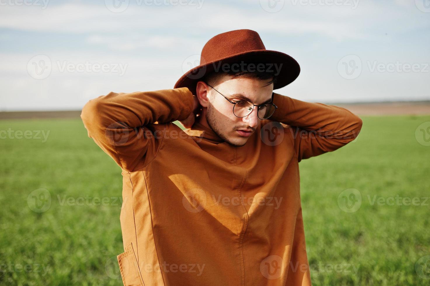 uomo alla moda con gli occhiali, giacca marrone e cappello in posa sul campo verde. foto