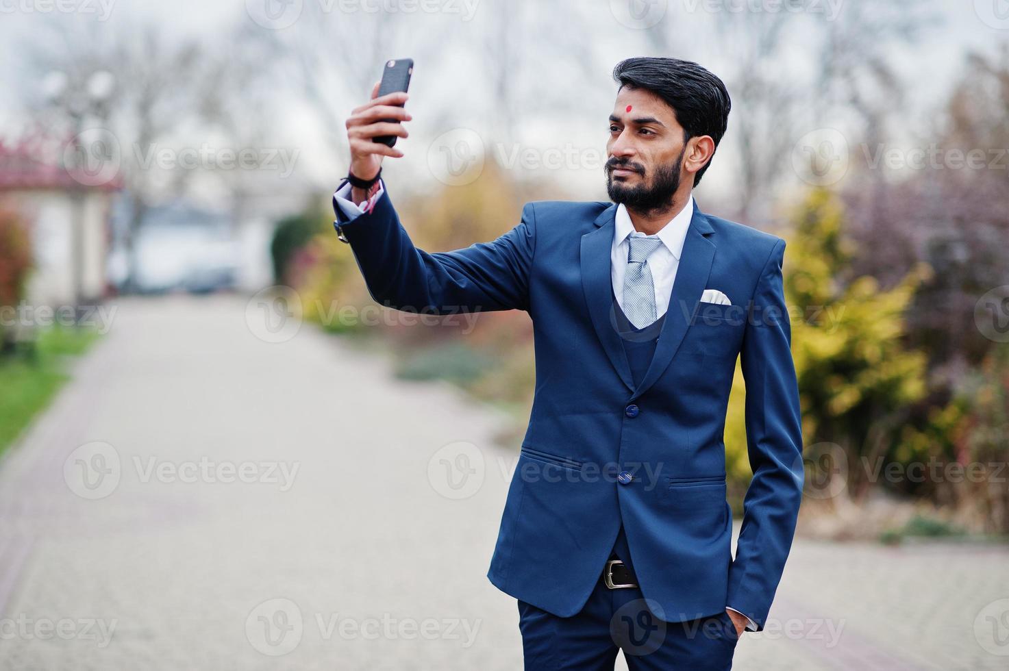 elegante barba indiano uomo d'affari con bindi sulla fronte, indossare su abito blu posato all'aperto facendo selfie al telefono cellulare. foto