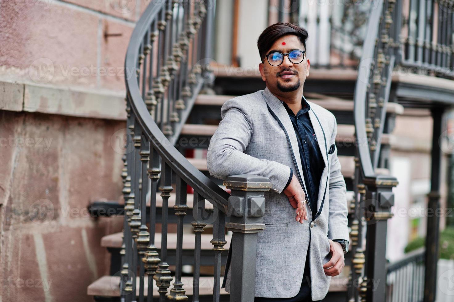 elegante uomo indiano con bindi sulla fronte e occhiali, indossa un abito posato all'aperto contro le scale di ferro. foto