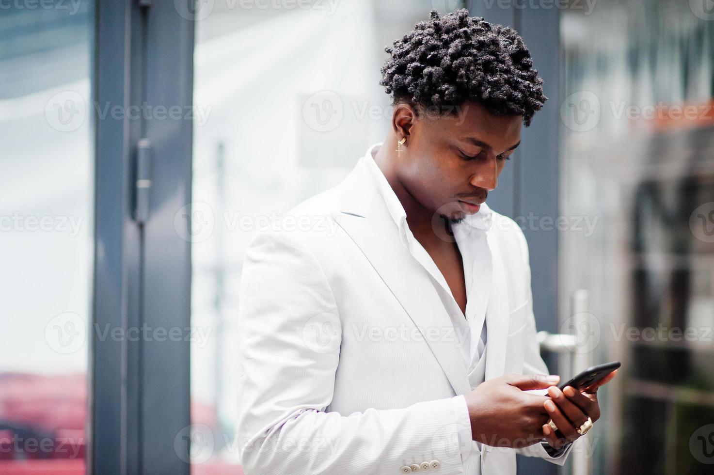 premuroso giovane gentiluomo afroamericano bello in abiti da cerimonia. nero elegante modello uomo in giacca bianca guardando il telefono cellulare. foto