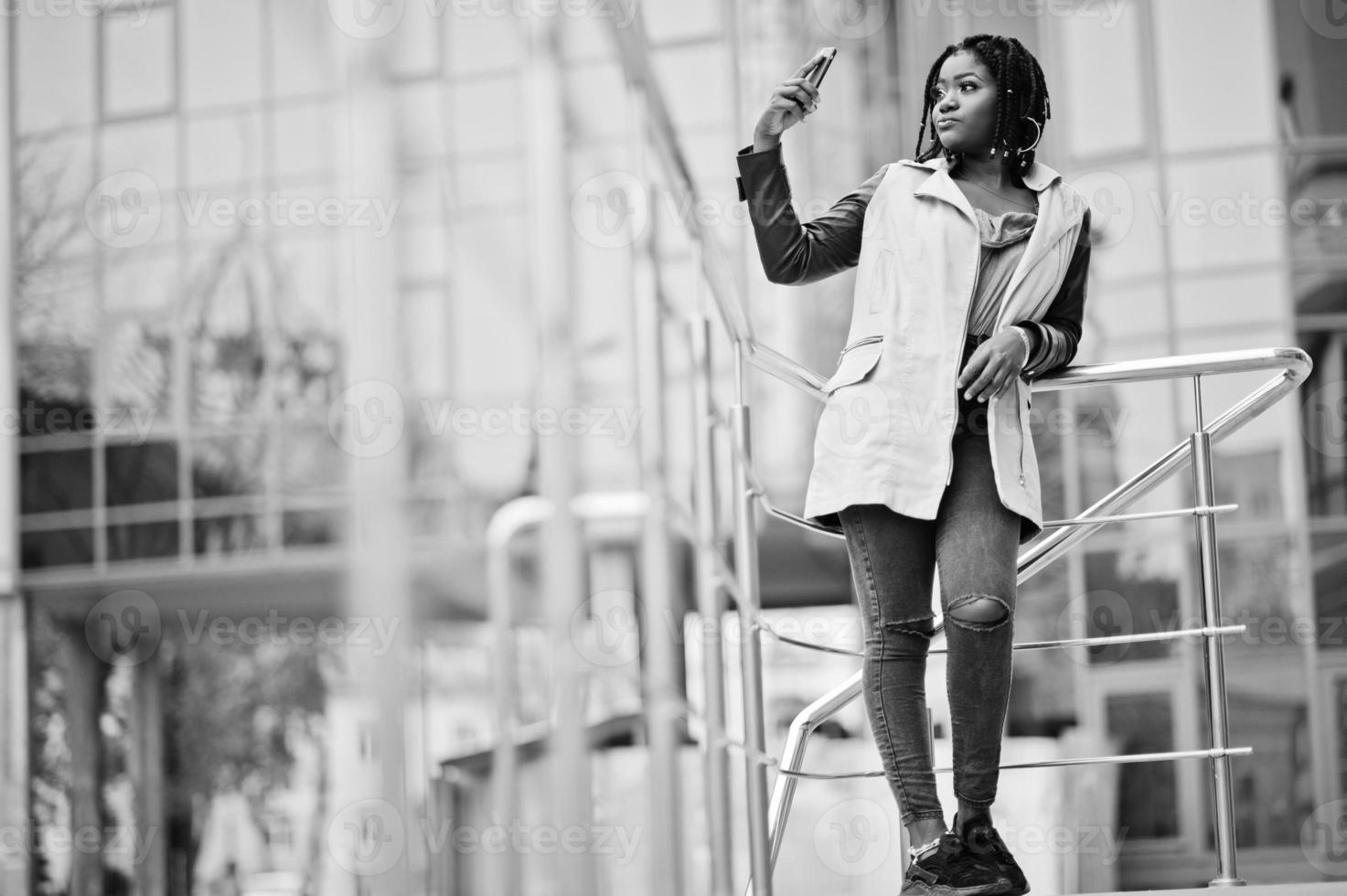 attraente donna afroamericana con dreadlocks in giacca posata vicino alle ringhiere contro un moderno edificio a più piani che fa selfie al telefono cellulare. foto