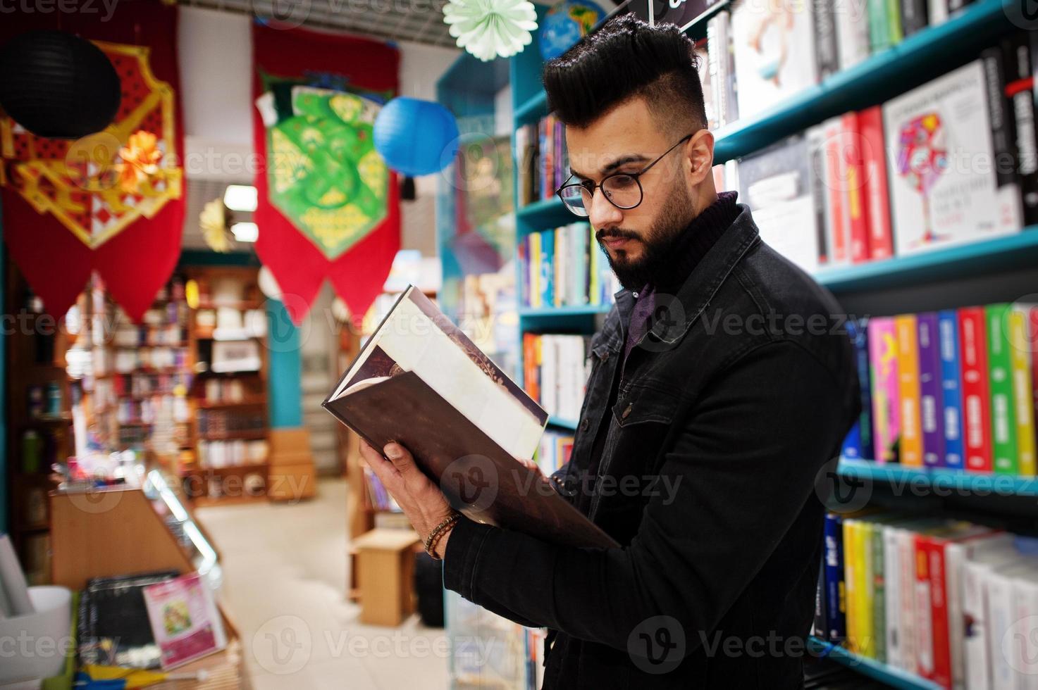 studente arabo alto e intelligente, indossa una giacca di jeans nera e occhiali da vista, in biblioteca con un libro a portata di mano. foto
