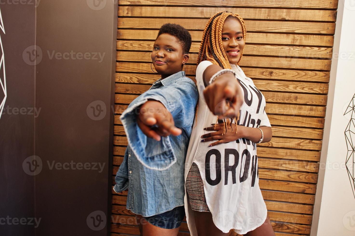 due donne africane in abiti casual eleganti che posano contro la parete di legno e mostrano le dita alla fotocamera. foto