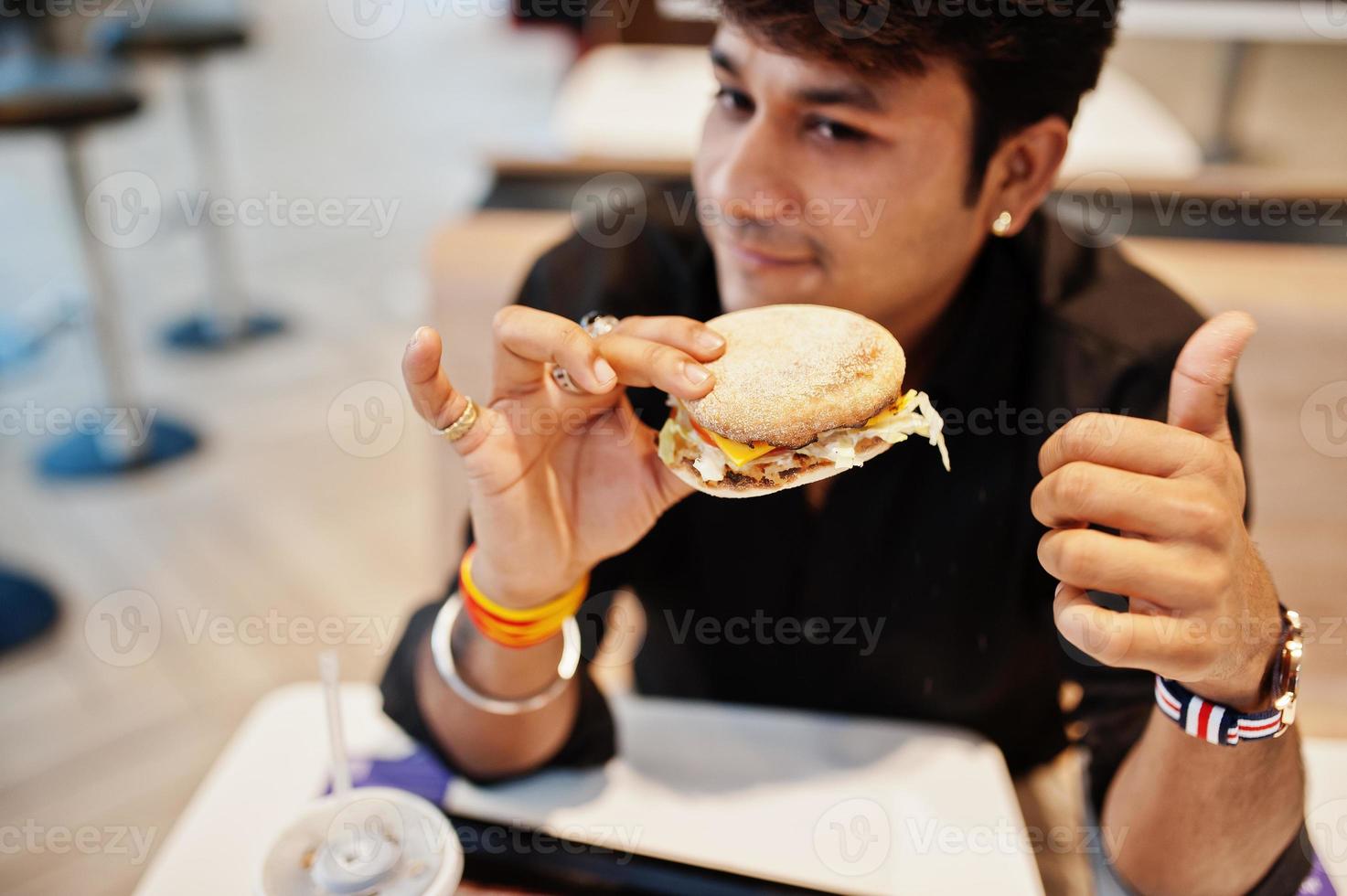 uomo indiano alla moda seduto al fast food cafe e mangiare hamburger. mostra il pollice in su. foto