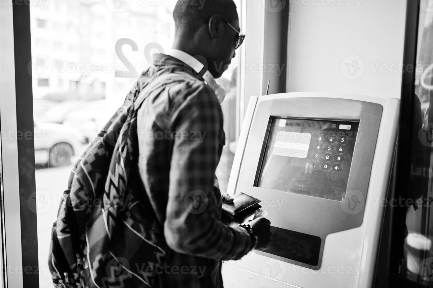 uomo afroamericano in camicia a scacchi con zaino che tiene il portafoglio in mano contro bancomat. viaggiatore nero. foto