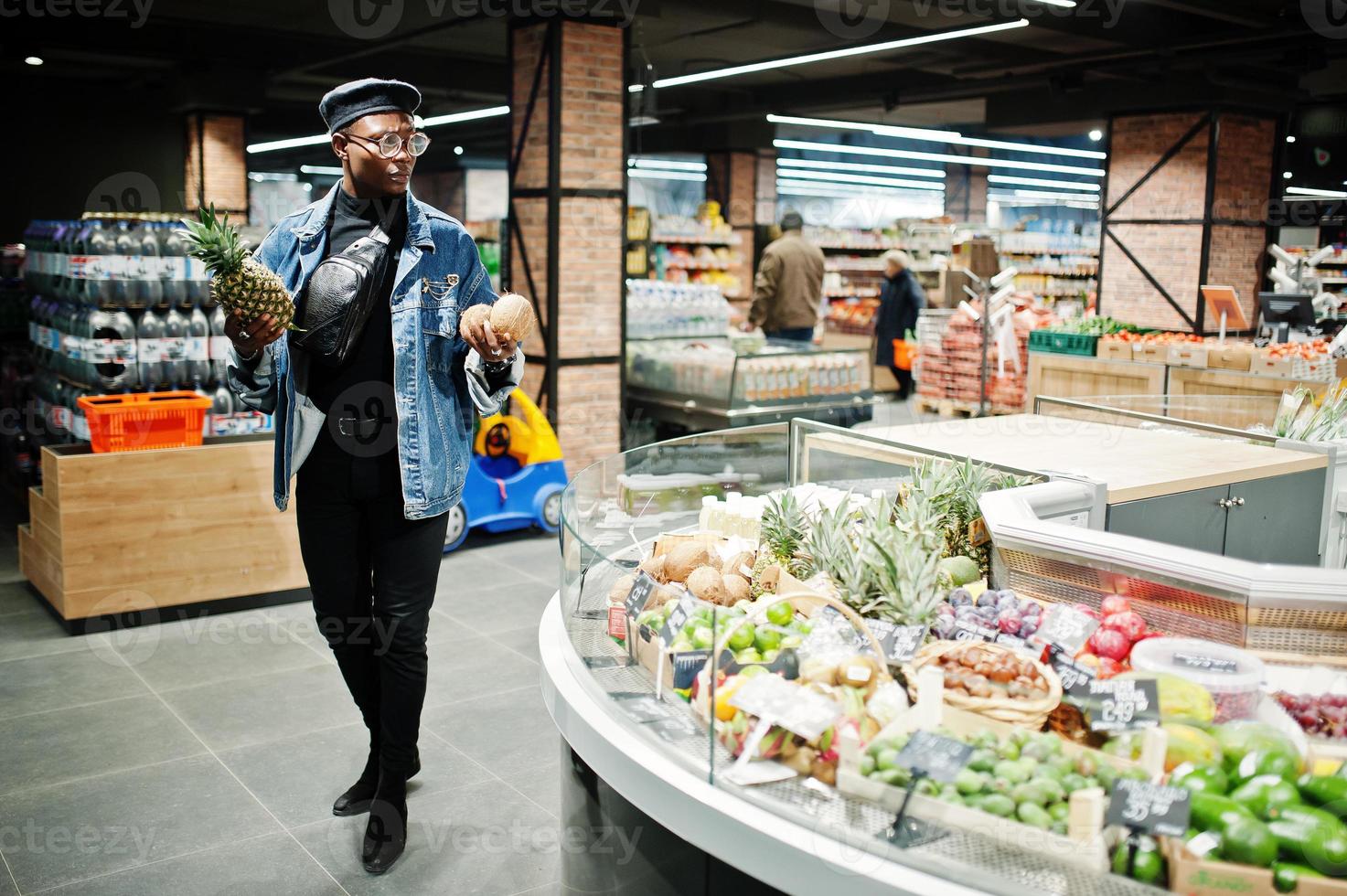 uomo afroamericano casual elegante alla giacca di jeans e berretto nero che tiene noci di cocco e ananas nella sezione di frutta biologica del supermercato. foto