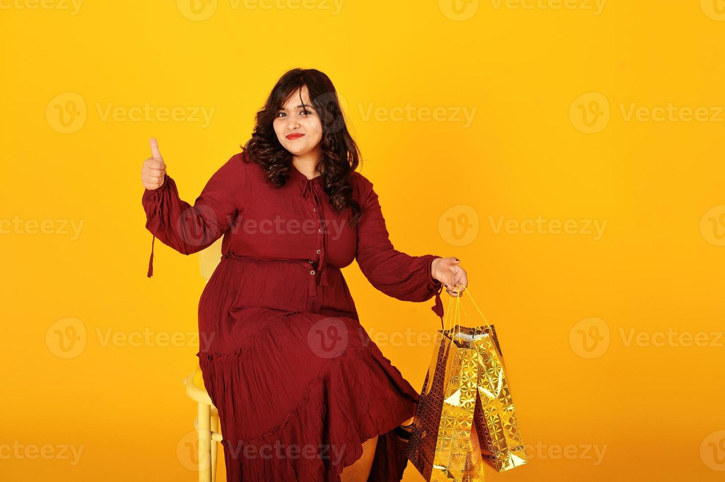 attraente donna del sud asiatico in abito rosso scuro posata in studio su sfondo giallo con borse della spesa dorate. foto