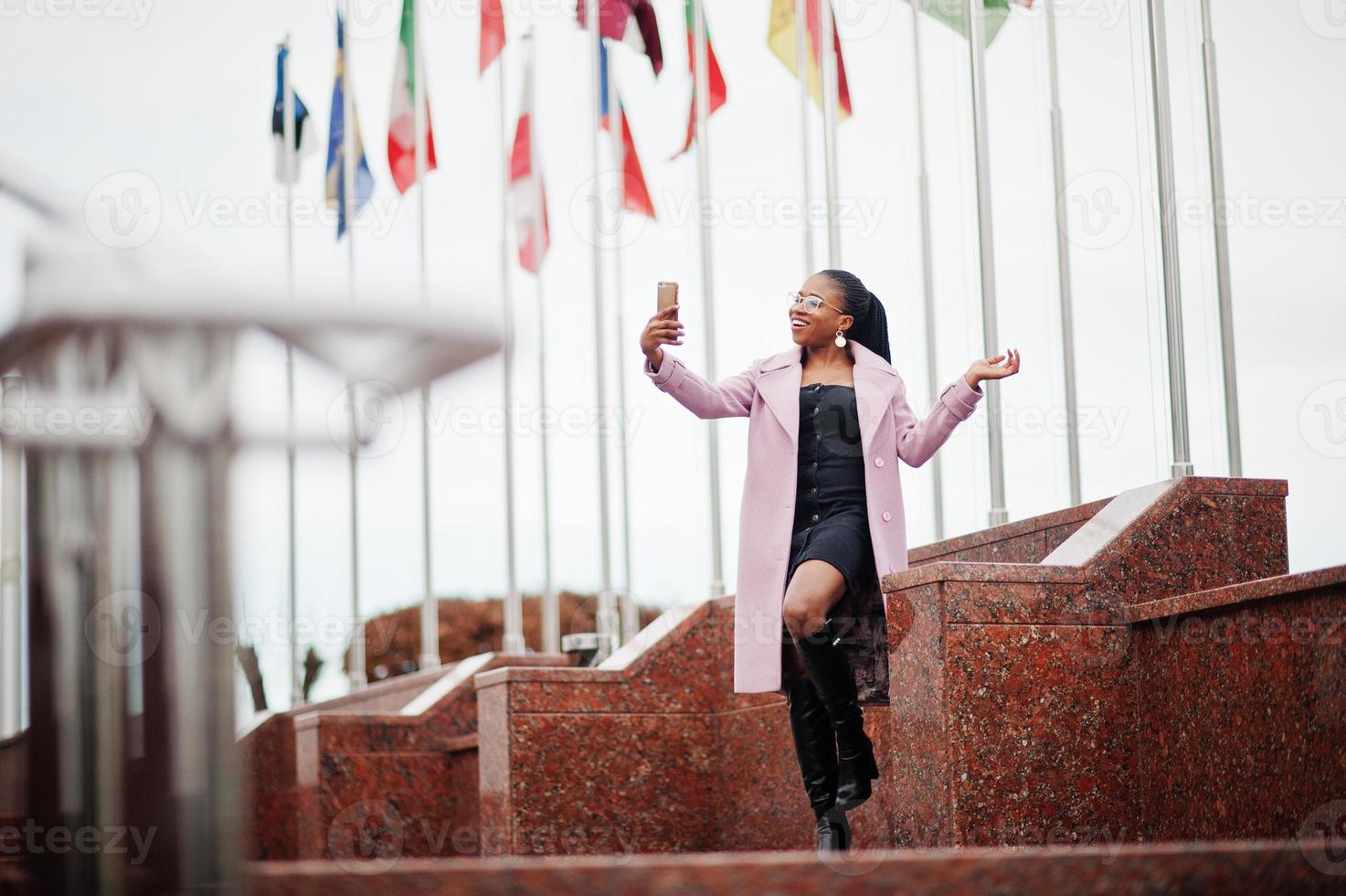 giovane ed elegante bella donna afroamericana in strada, indossando un cappotto di moda, contro le bandiere di diversi paesi del mondo, con il telefono cellulare a portata di mano. foto