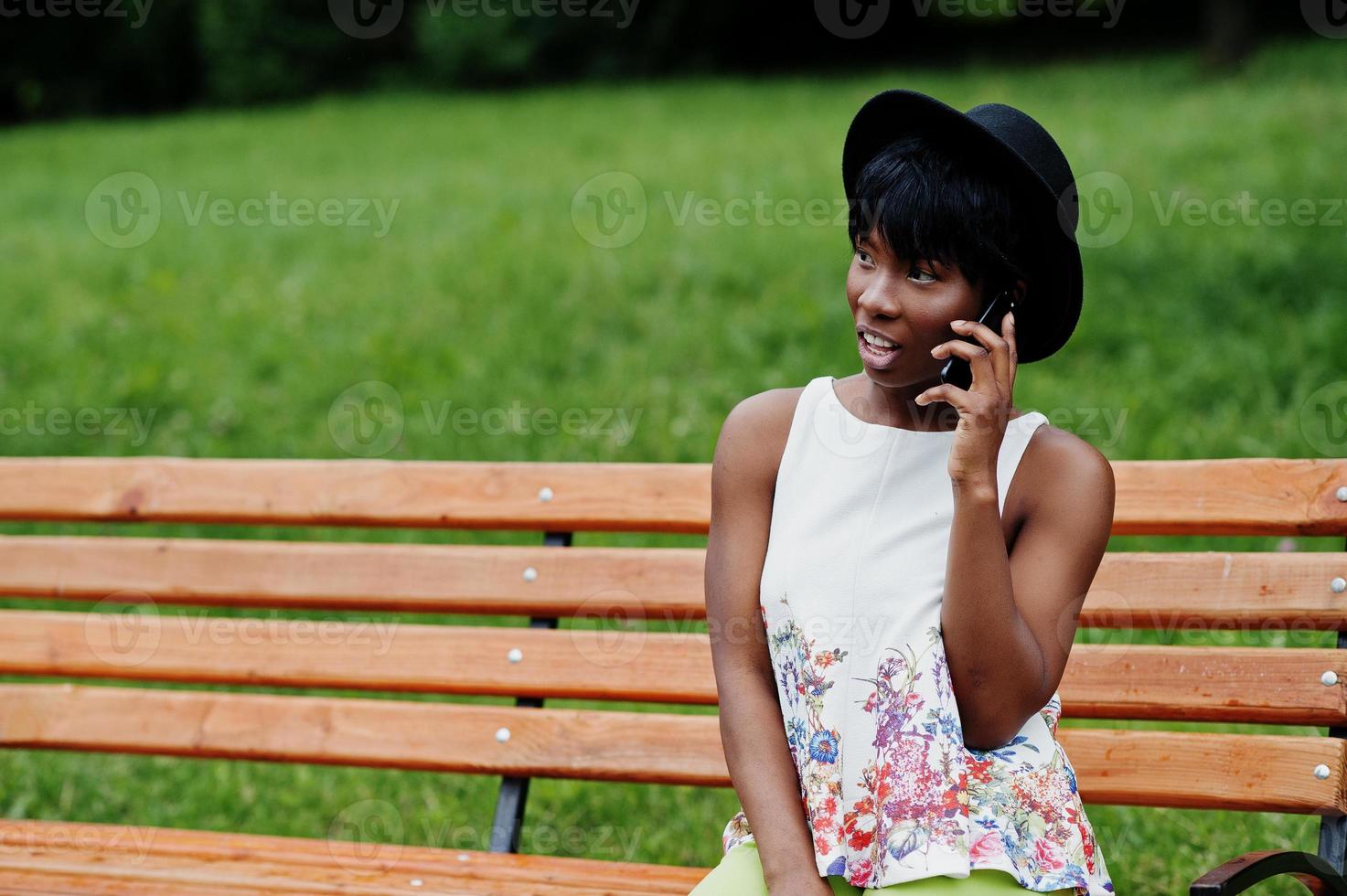 incredibile donna modello afroamericano in pantaloni verdi e cappello nero in posa su una panchina al parco e parlando al telefono cellulare. foto