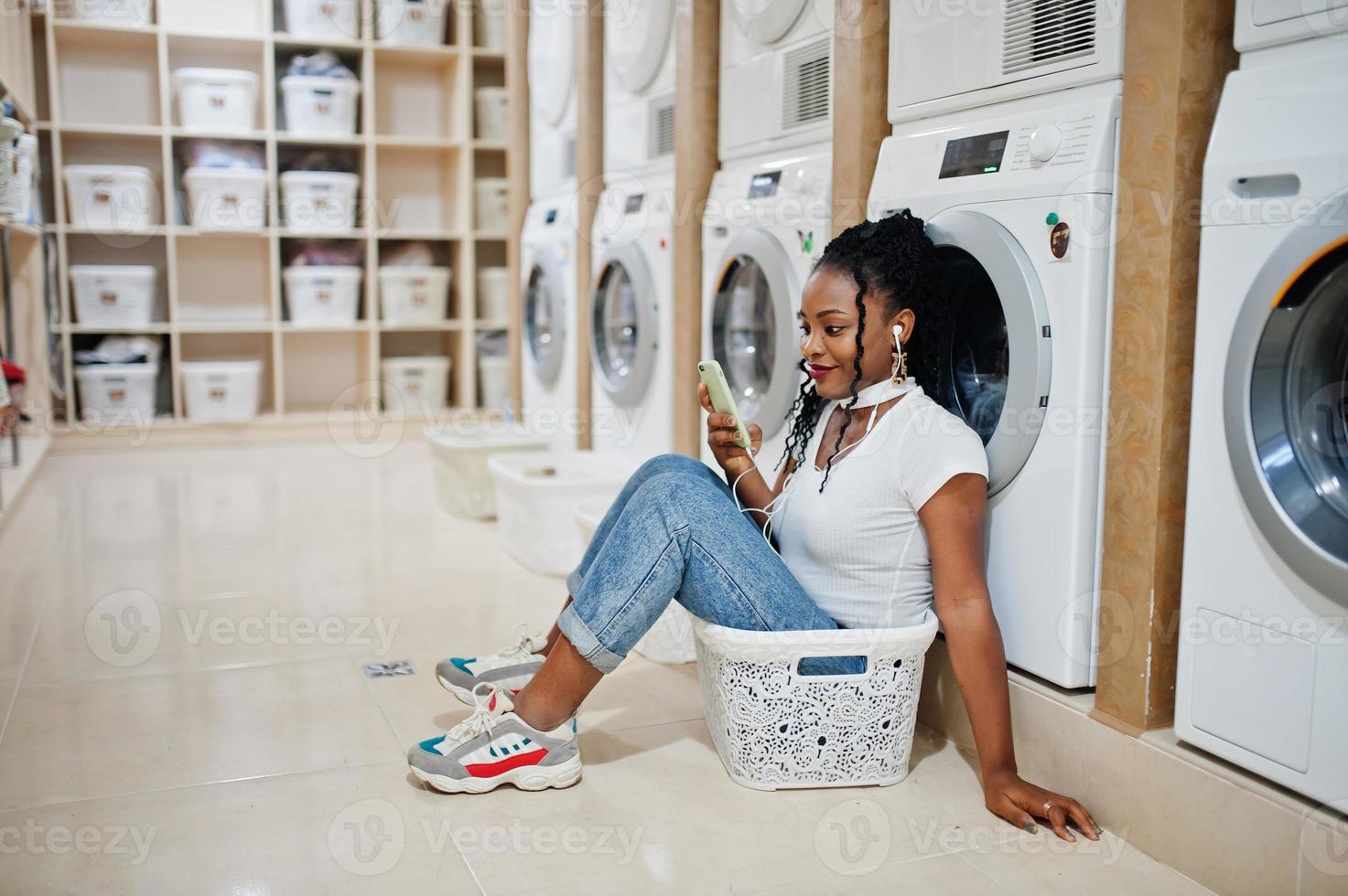 allegra donna afroamericana vicino alla lavatrice ascoltando musica con gli auricolari dal telefono cellulare nella lavanderia self-service. foto