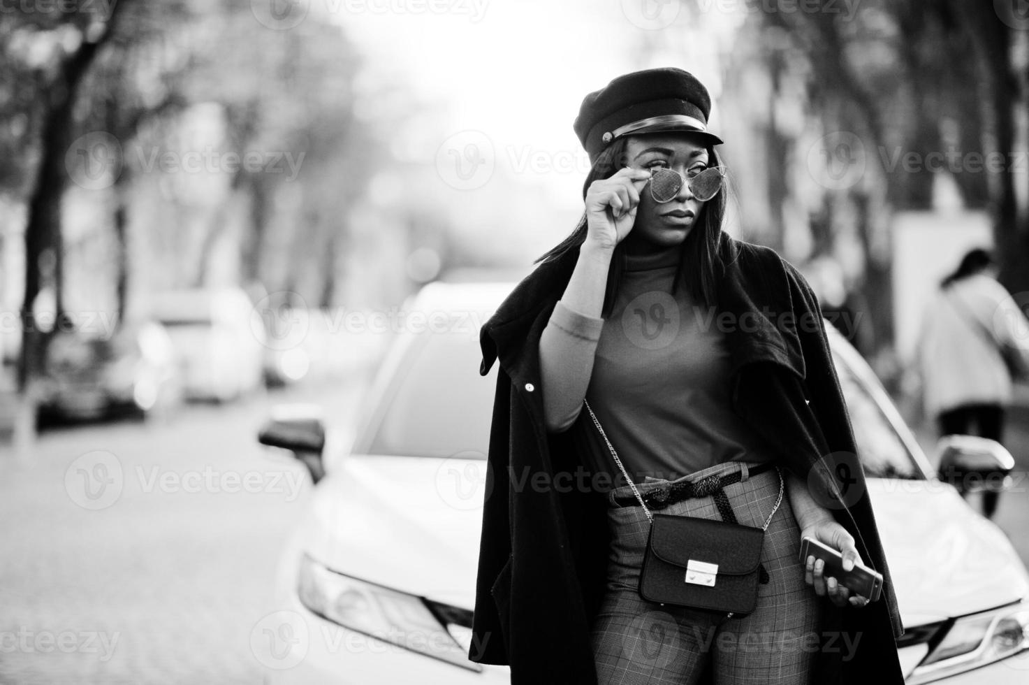 ragazza di moda afroamericana in cappotto, berretto da strillone e occhiali da sole poste in strada contro un'auto bianca da lavoro. foto