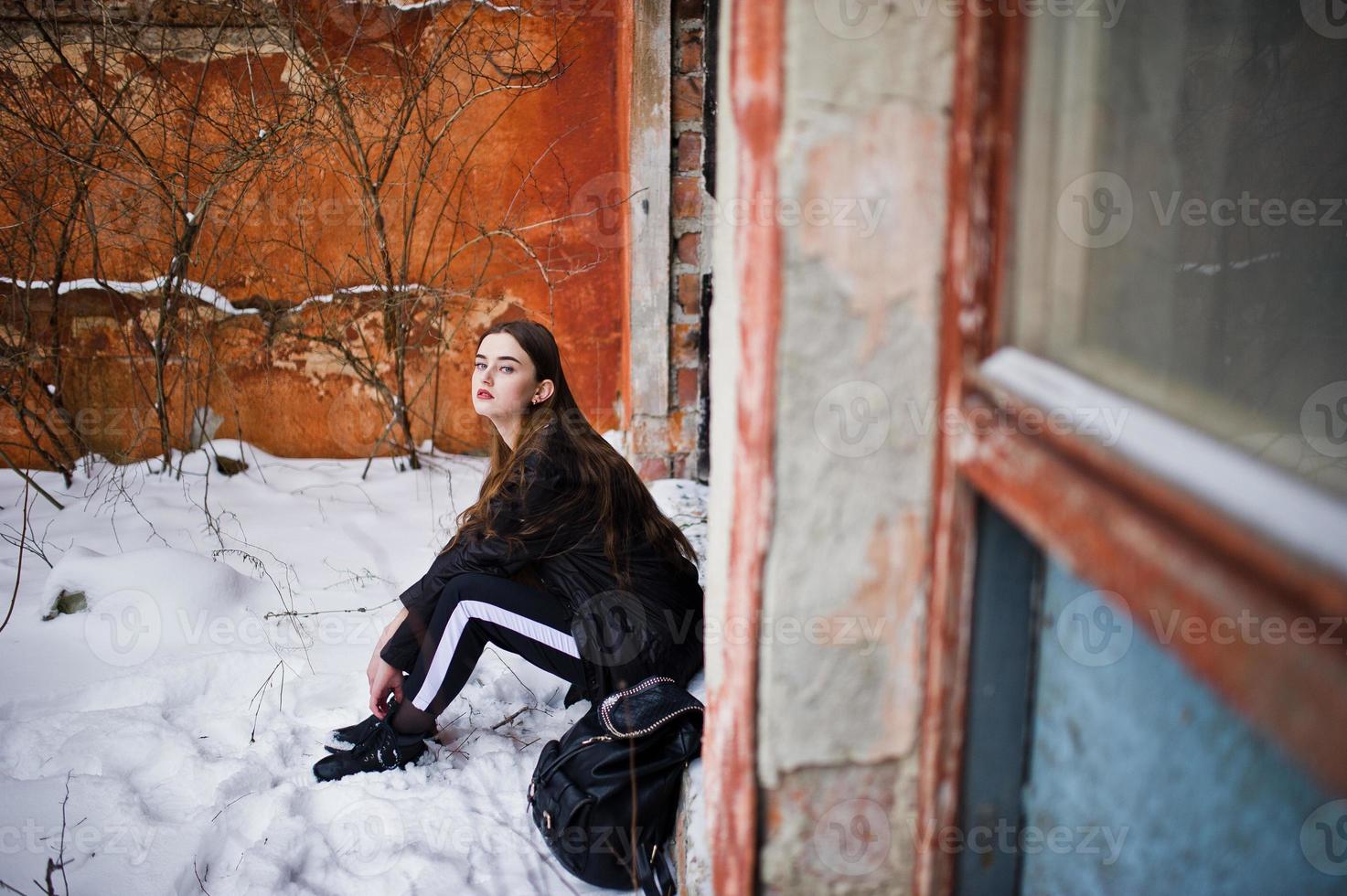 modello bruna alla moda gambe lunghe in lungo mantello nero poste all'aperto al giorno d'inverno contro il vecchio muro di lerciume. foto