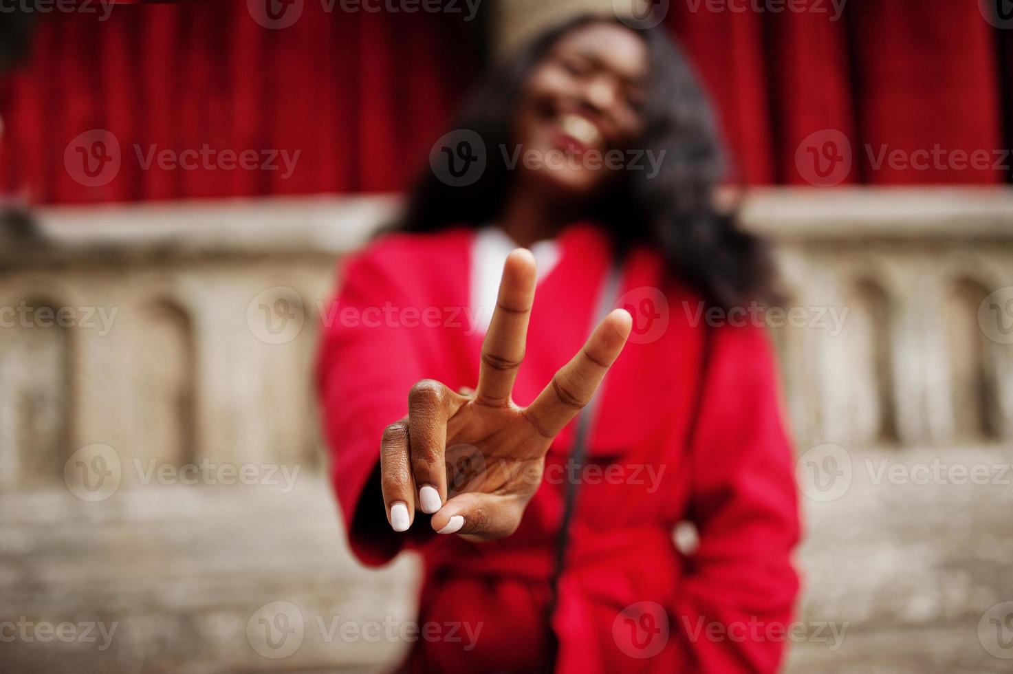 elegante donna afroamericana in cappotto rosso posata contro vecchie colonne e mostra il segno di due dita. foto