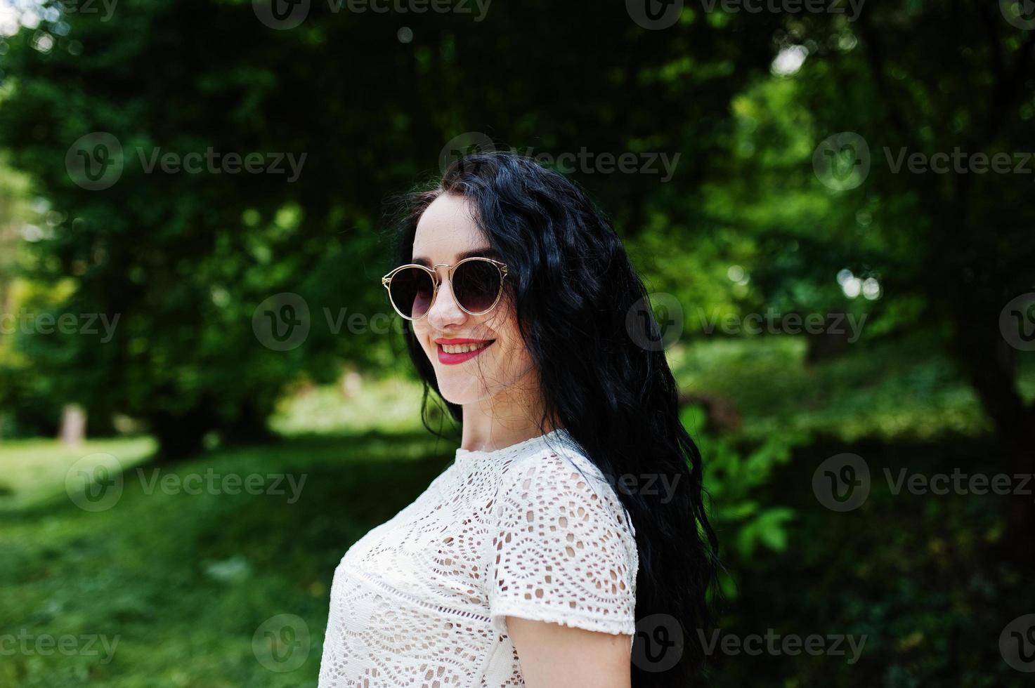 ragazza bruna in camicetta bianca con occhiali da sole poste al parco. foto