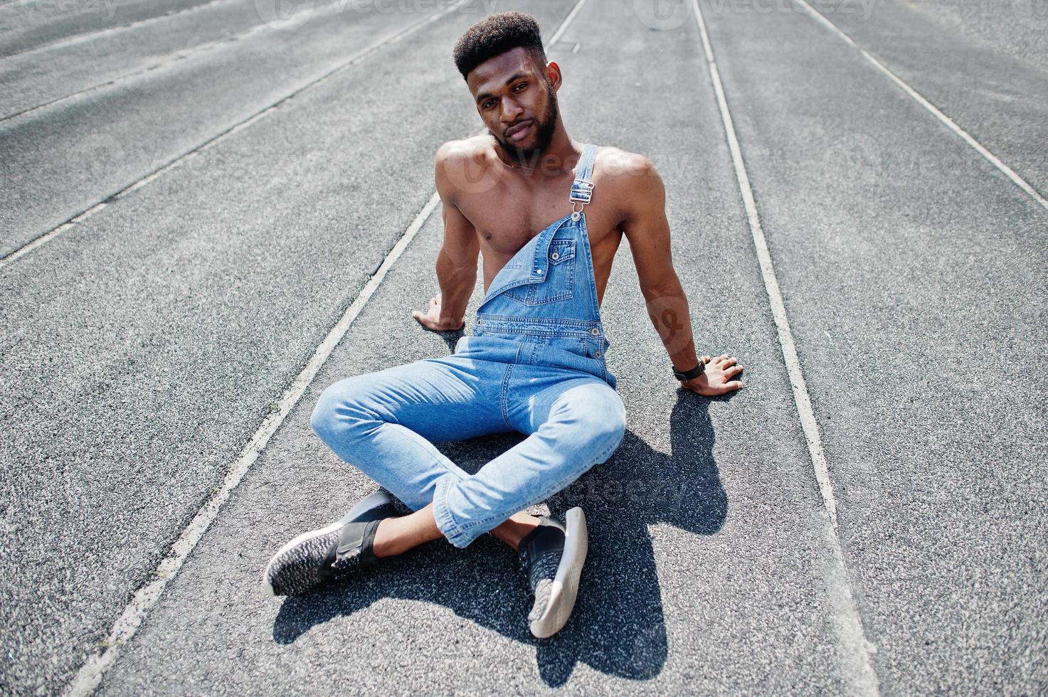 bell'uomo a torso nudo afroamericano sexy in tuta di jeans seduto all'ippodromo dello stadio. ritratto di uomo nero alla moda. foto