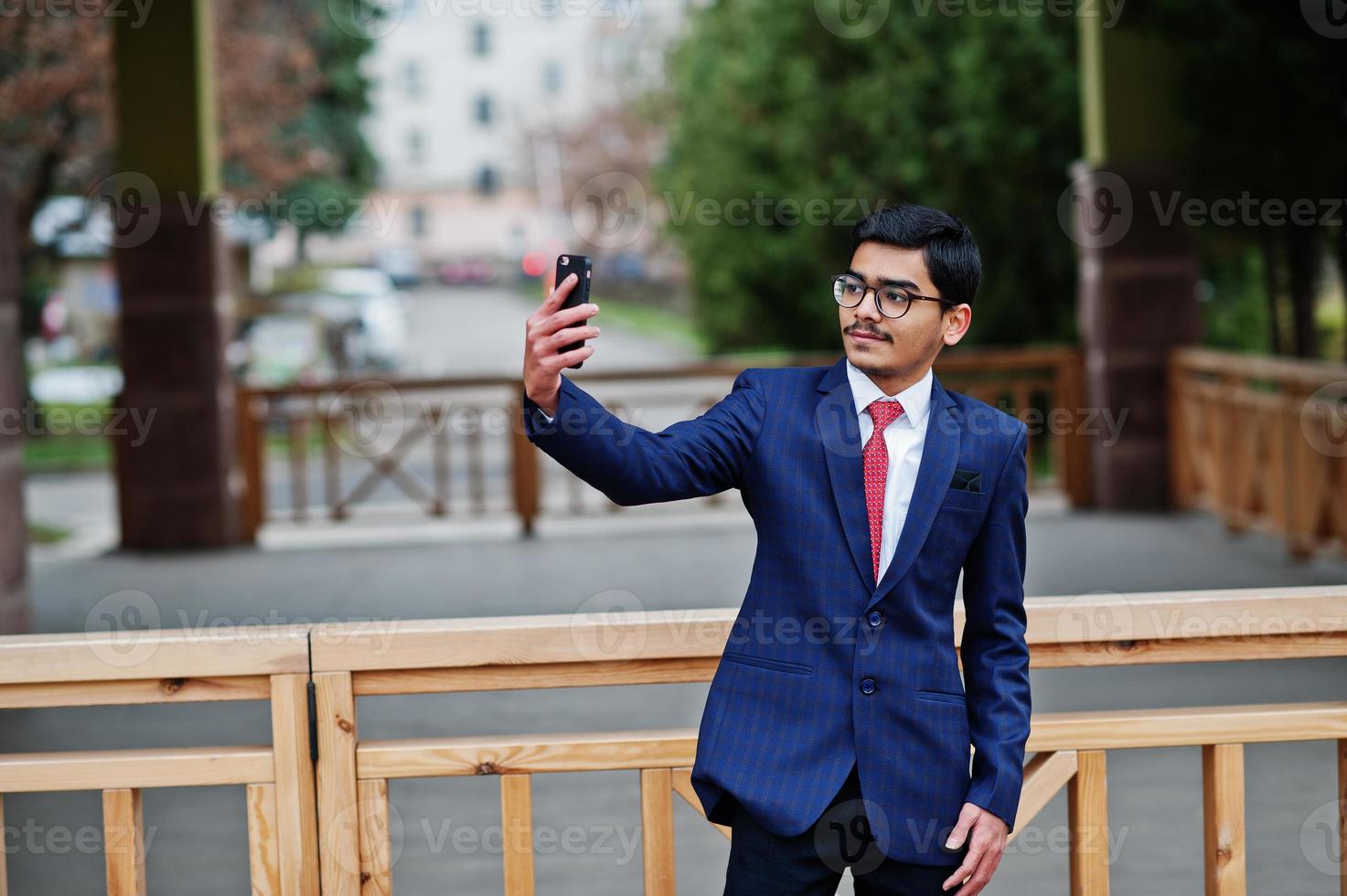 giovane indiano con gli occhiali, indossare un abito con cravatta rossa in posa all'aperto e fare selfie sul cellulare. foto