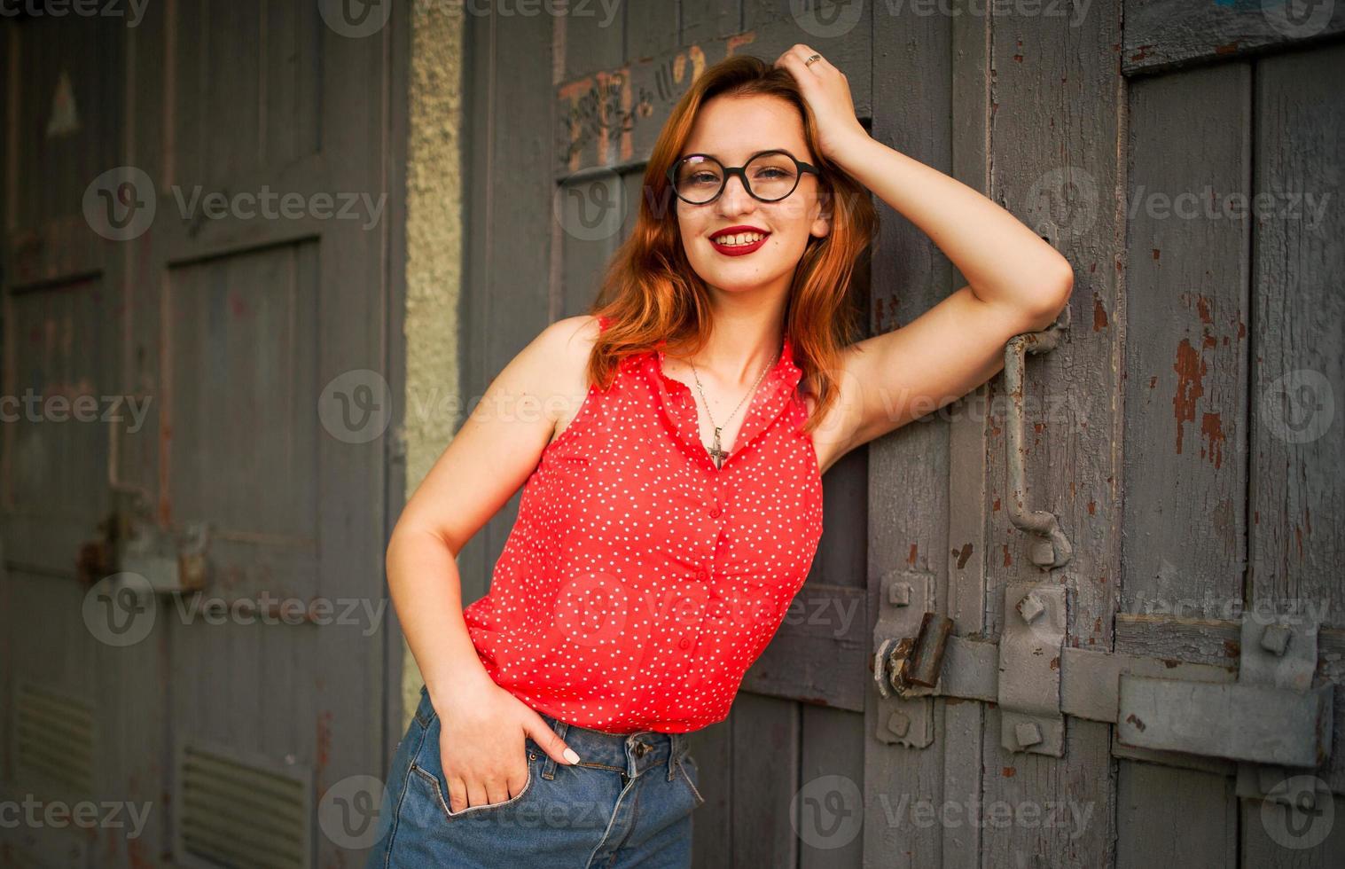 attraente donna dai capelli rossi in occhiali, indossare su camicetta rossa e gonna di jeans in posa. foto