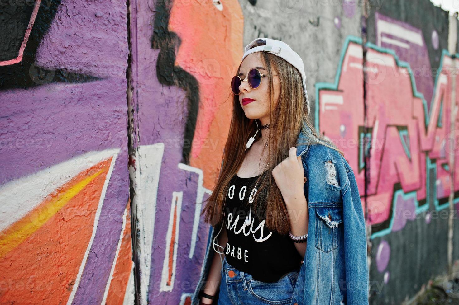 elegante ragazza hipster casual in berretto, occhiali da sole e jeans indossano, ascoltando musica dalle cuffie del telefono cellulare contro il grande muro di graffiti. foto