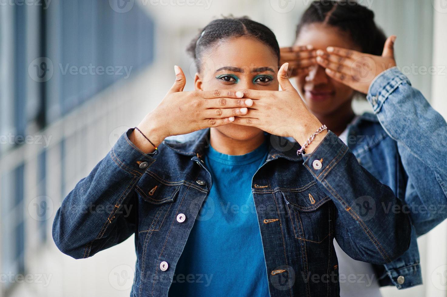 due amiche africane in giacca di jeans coprirono la bocca e gli occhi con le mani al coperto insieme. foto