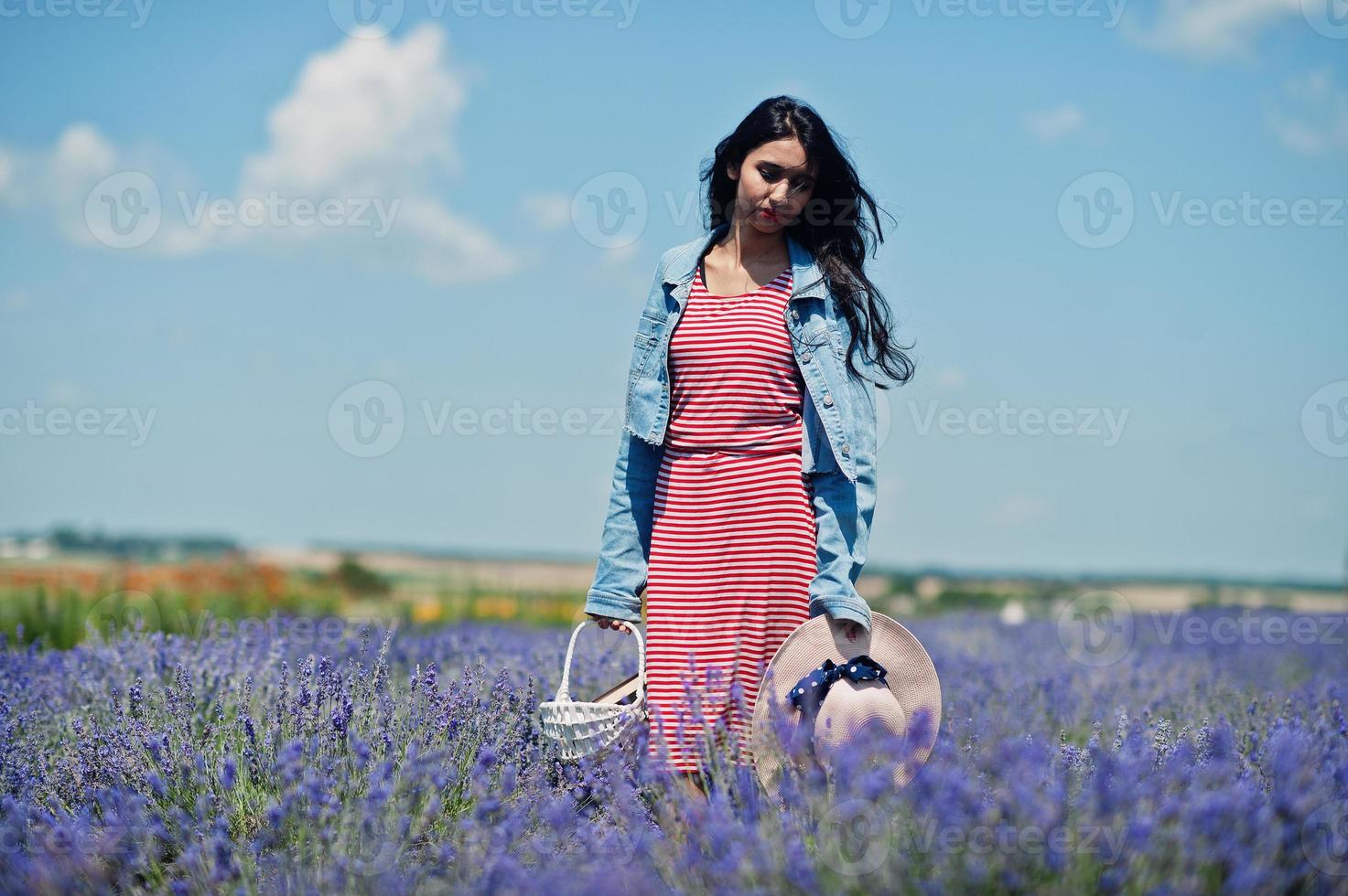bella ragazza indiana in abito estivo e giacca di jeans in campo di lavanda viola con cesto in mano e cappello. foto