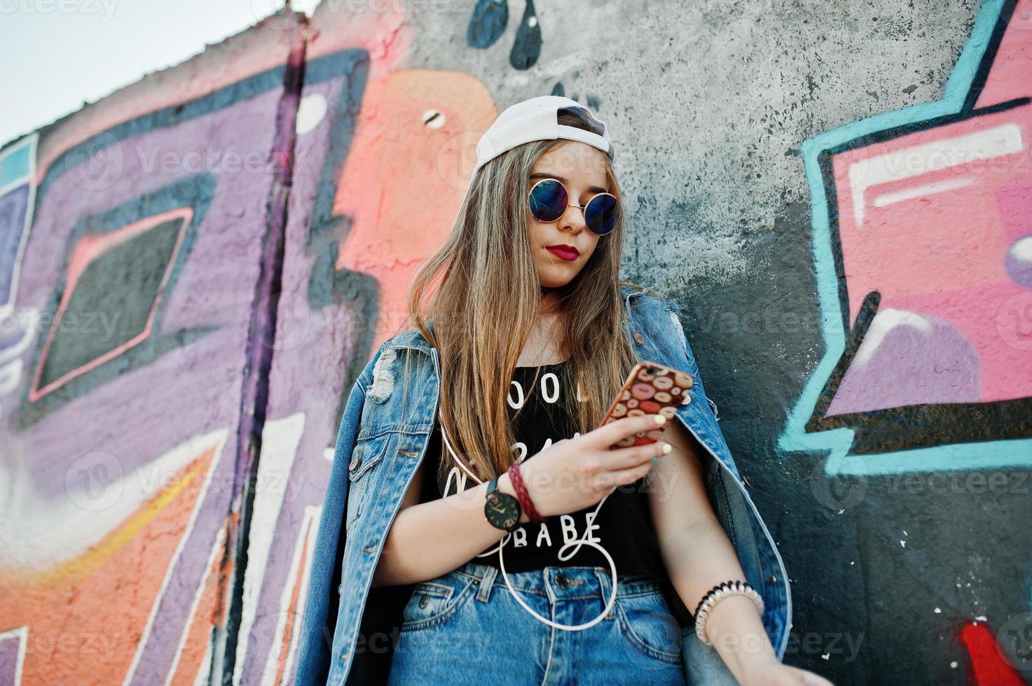 elegante ragazza hipster casual in berretto, occhiali da sole e jeans indossano, ascoltando musica dalle cuffie del telefono cellulare contro il grande muro di graffiti. foto