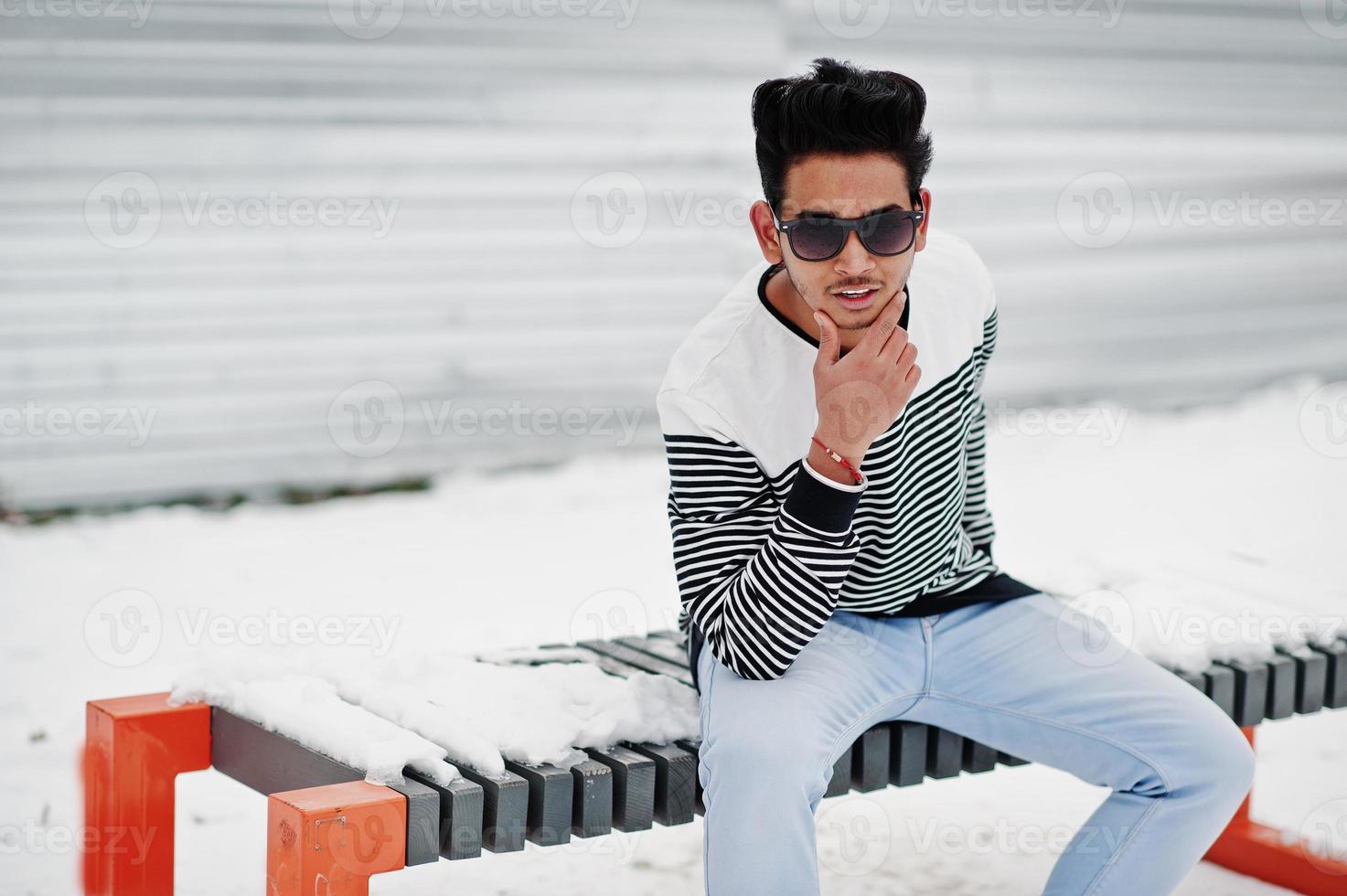 giovane indiano casual in occhiali da sole posato in una giornata invernale e seduto su una panchina. foto