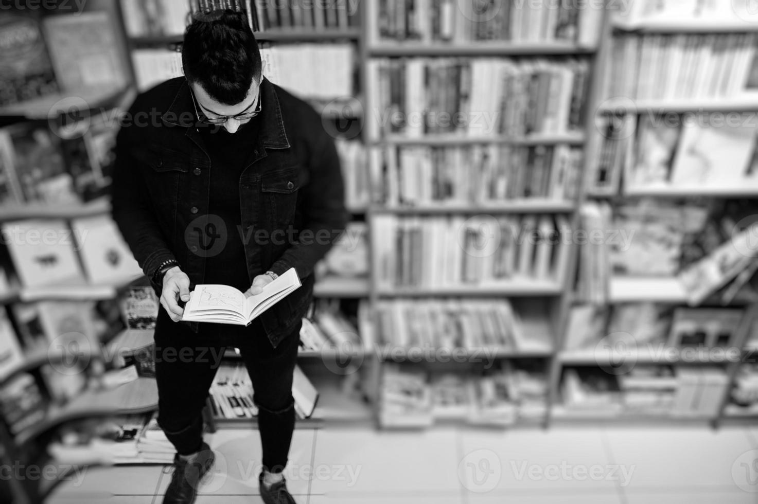 studente arabo alto e intelligente, indossa una giacca di jeans nera e occhiali da vista, in biblioteca con un libro a portata di mano. foto