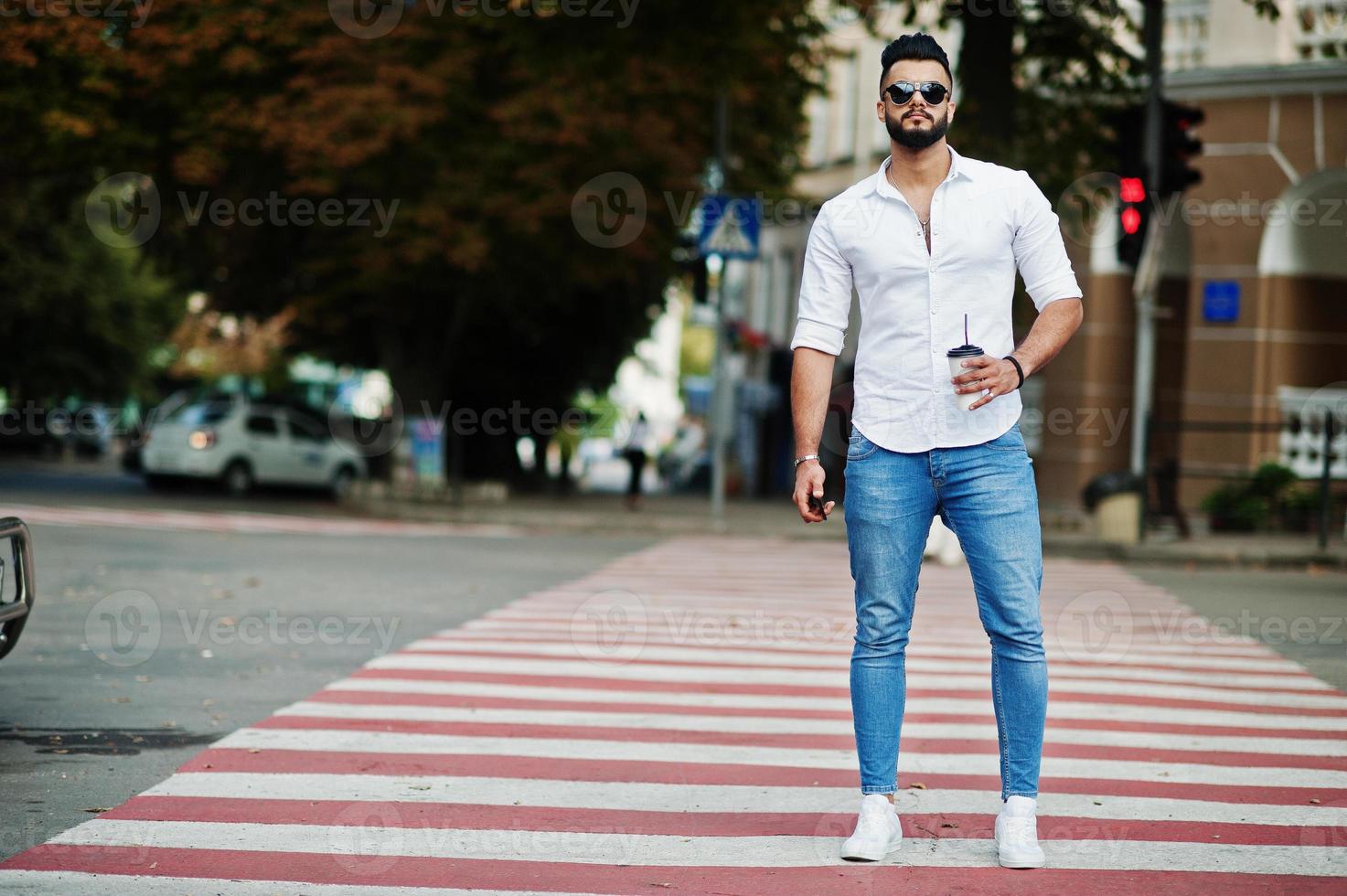 elegante uomo arabo alto modello in camicia bianca, jeans e occhiali da sole posato in una strada della città. barba attraente ragazzo arabo con una tazza di caffè che cammina sul passaggio pedonale o sul passaggio pedonale. foto