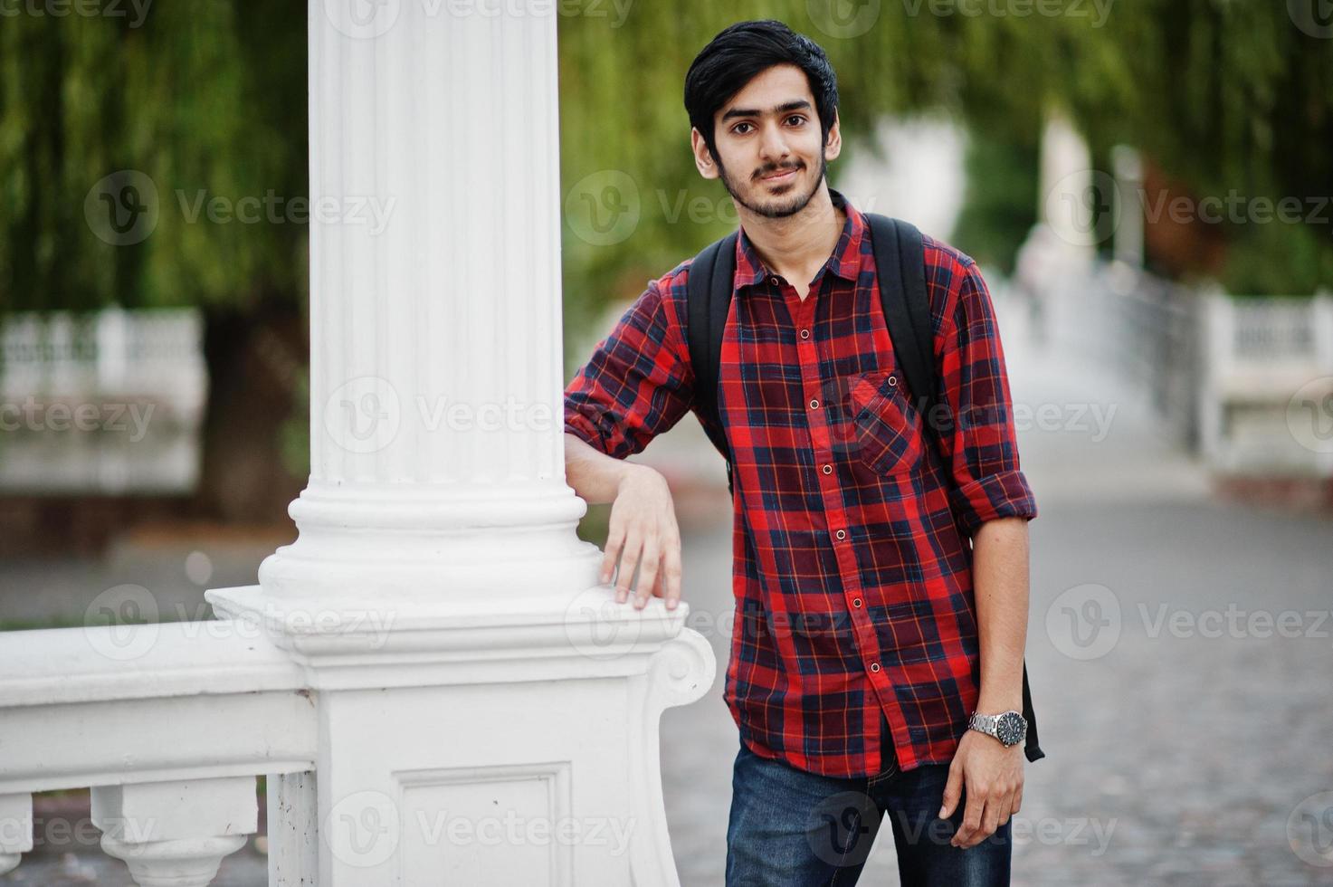 giovane studente indiano in camicia a scacchi e jeans con zaino in posa sulla città di sera contro la colonna. foto