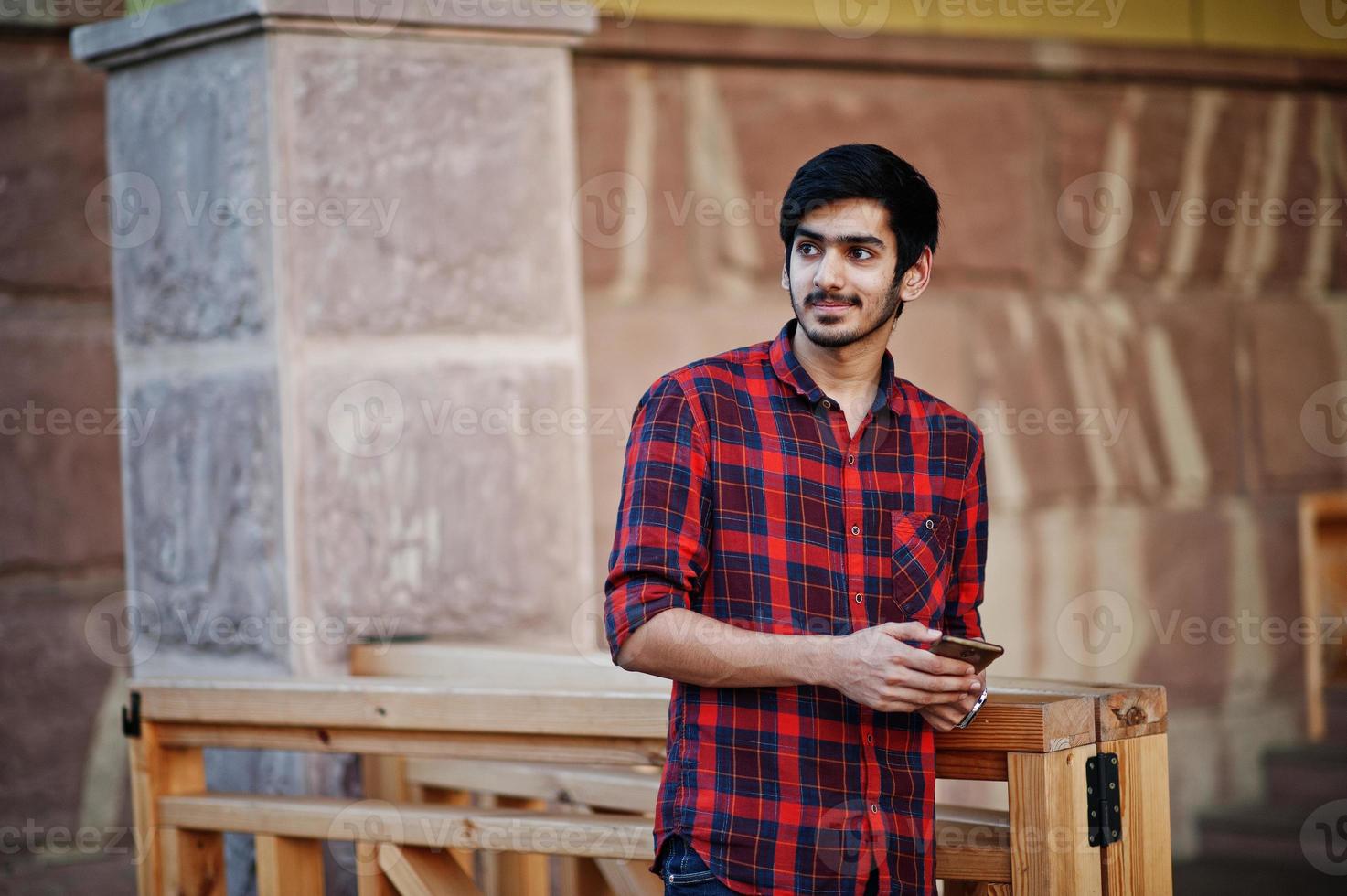 giovane studente indiano in camicia a scacchi rossa e jeans posato in città con il cellulare a portata di mano. foto