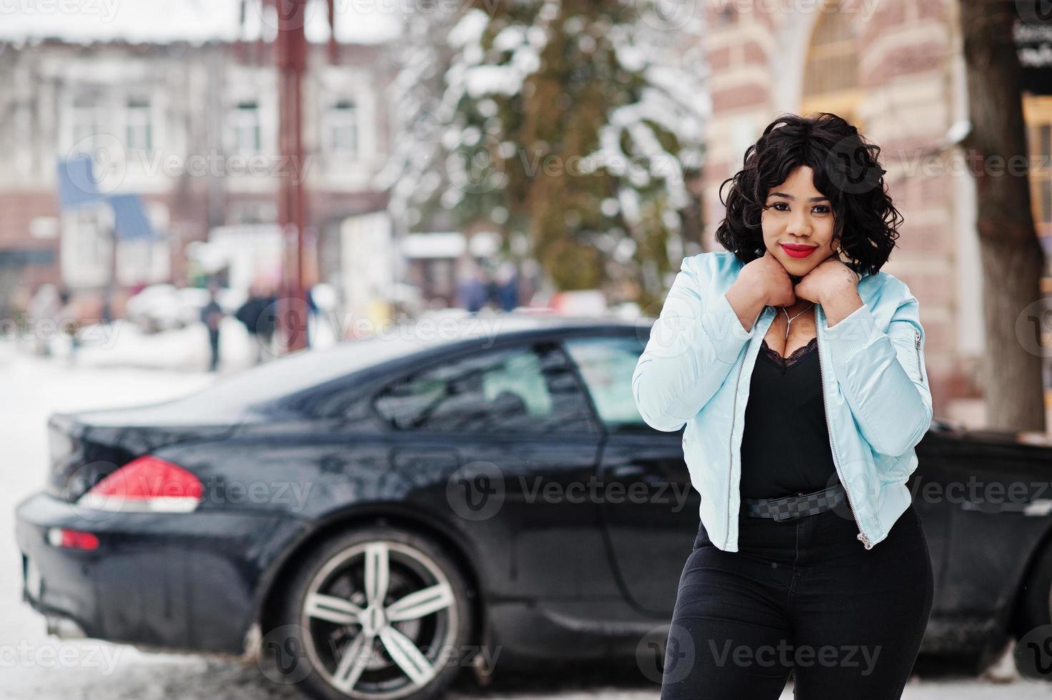 elegante modello afroamericano plus size con il telefono cellulare a portata di mano durante la giornata invernale nevosa contro l'auto nera. foto