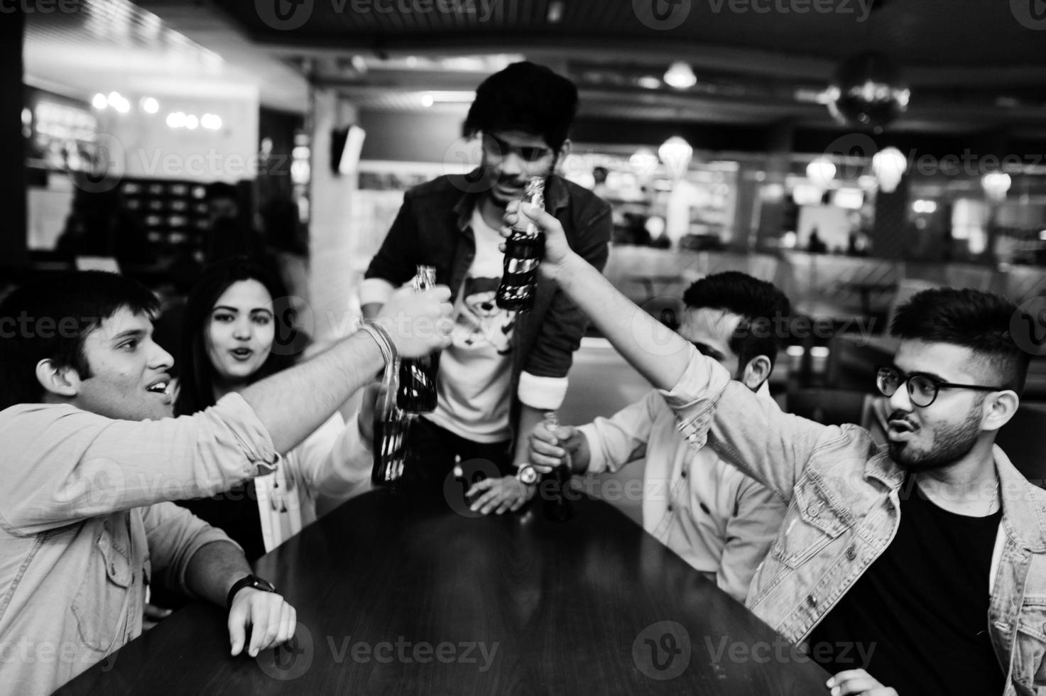 gruppo di amici asiatici alla moda indossa jeans seduti a tavola e tifa con bottiglie di soda nel club. foto