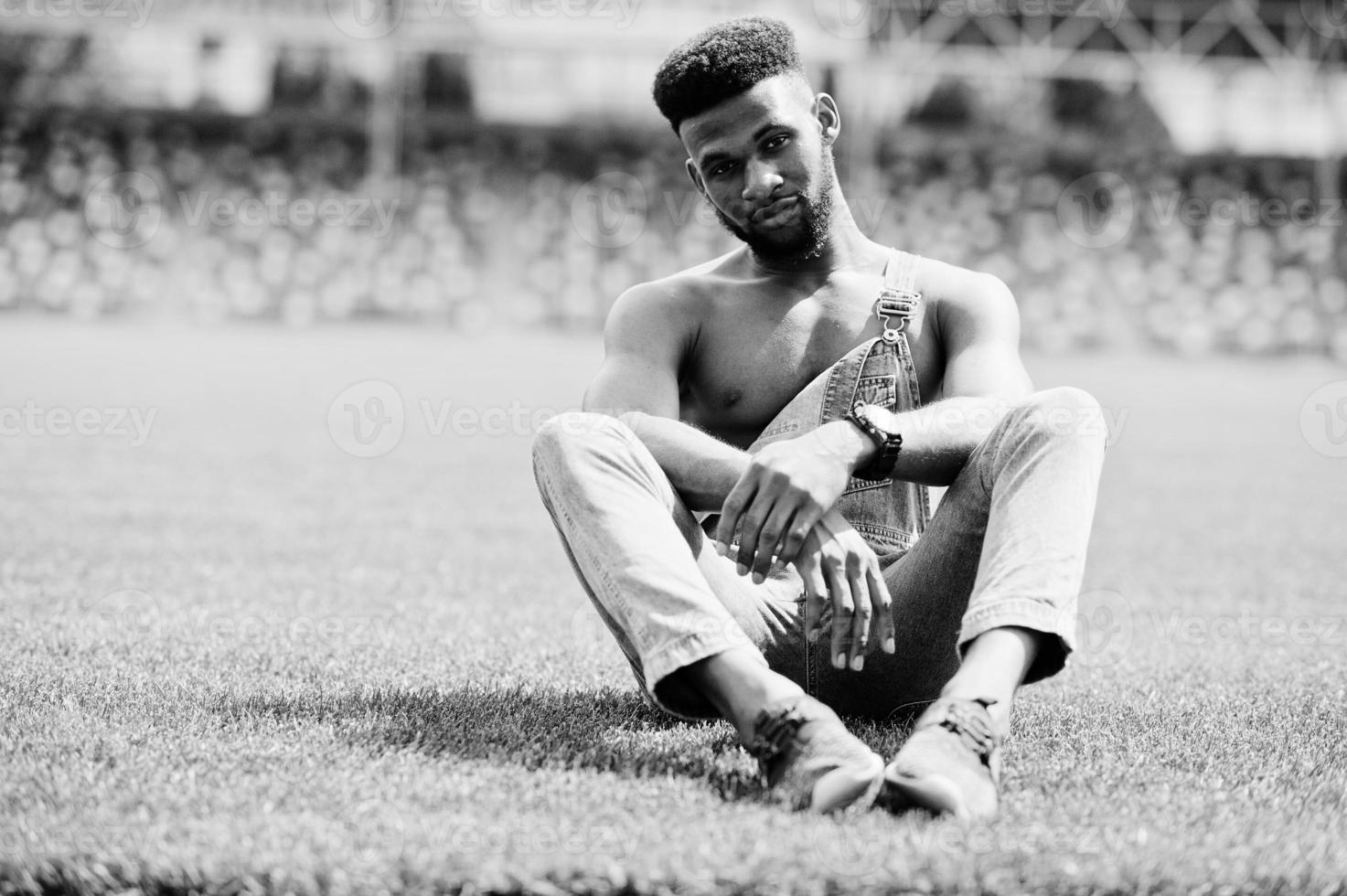 bell'uomo a torso nudo afroamericano sexy in tuta di jeans seduto all'erba verde del campo di calcio dello stadio. ritratto di uomo nero alla moda. foto