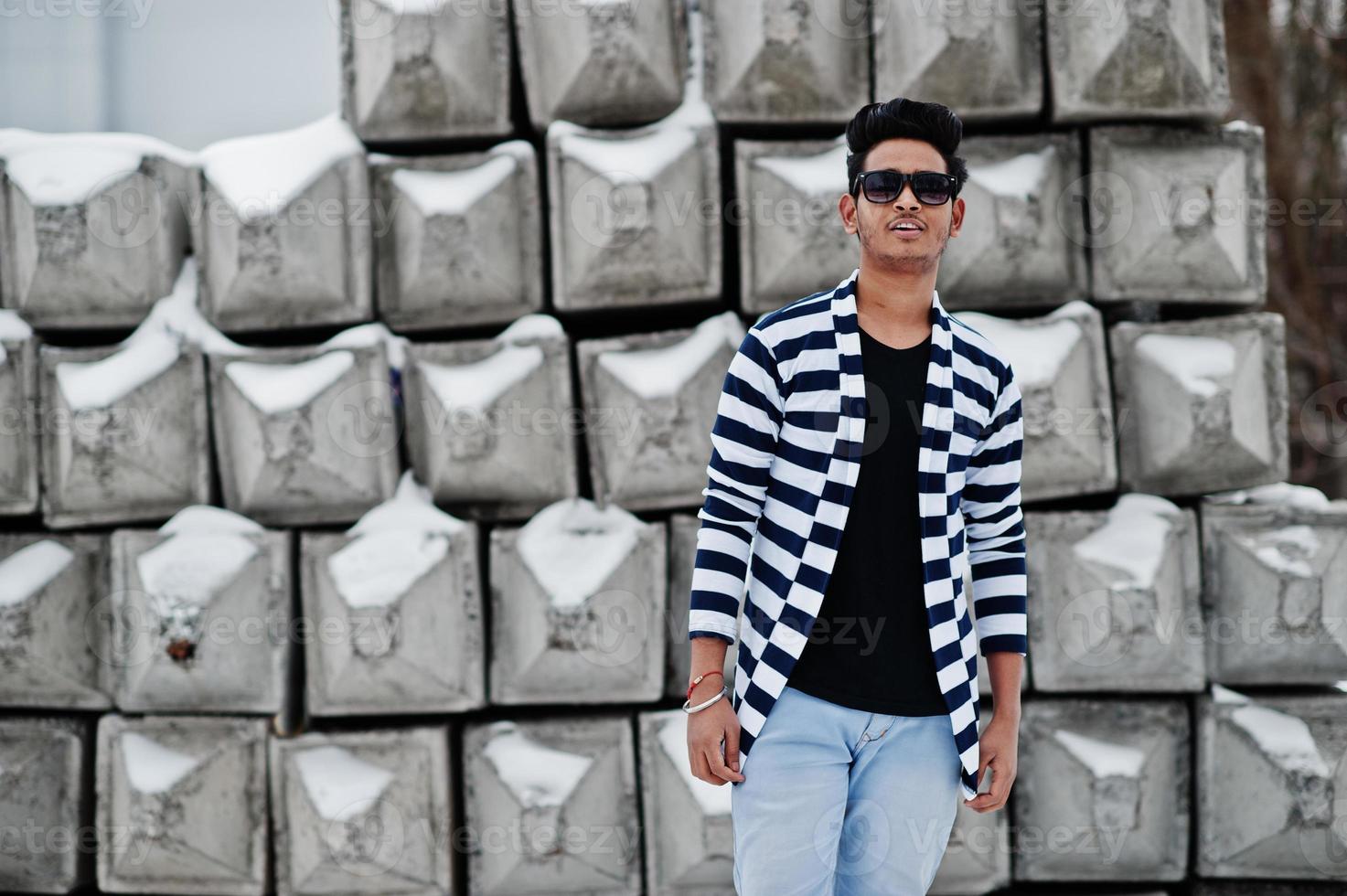 giovane indiano casual in maglione spogliato e occhiali da sole posato contro blocchi di pietra. foto