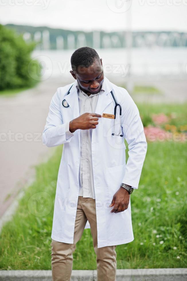 giovane medico maschio afroamericano in camice bianco con uno stetoscopio posato all'aperto. foto