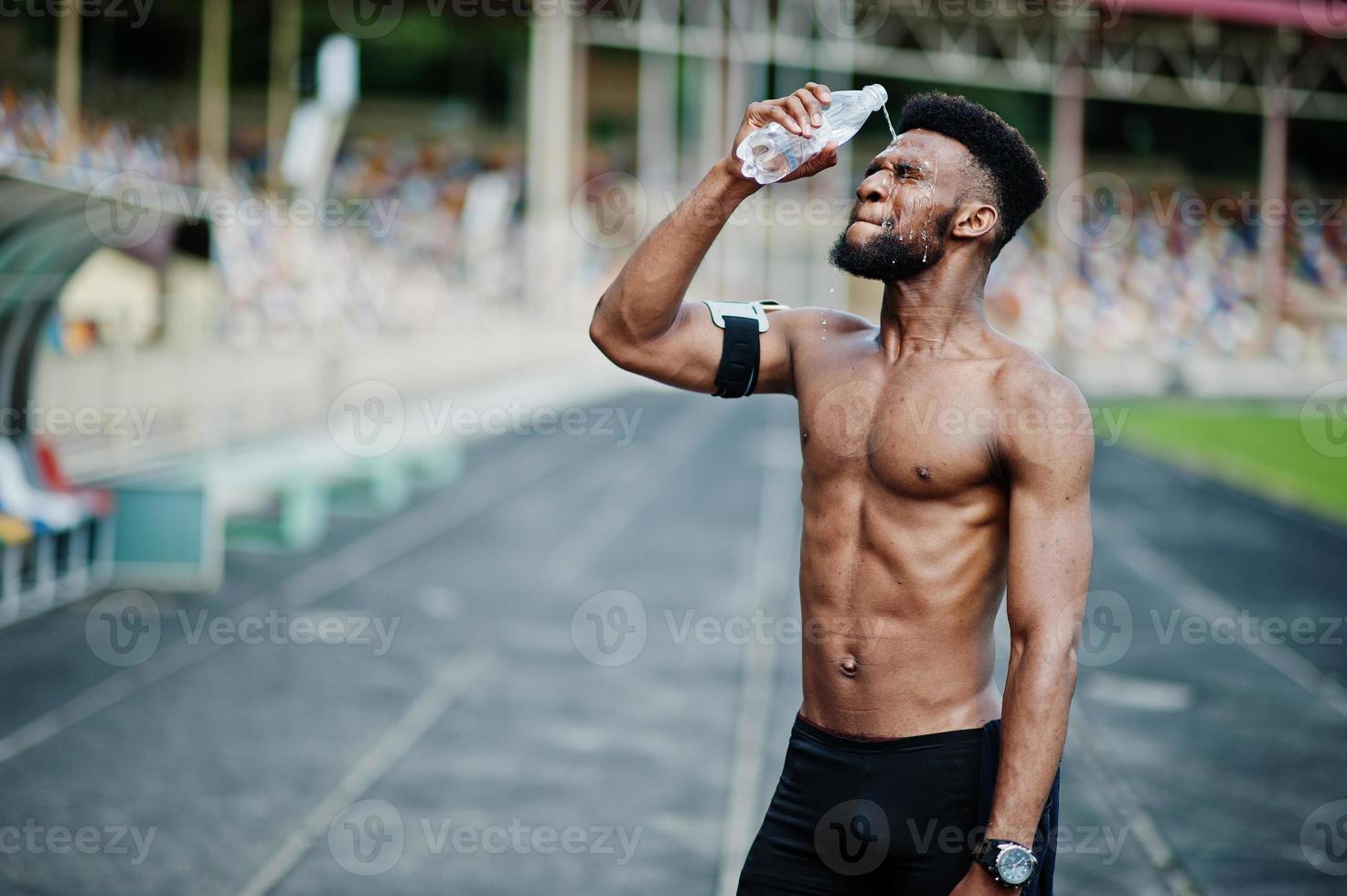 atleta maschio afroamericano sport sexy torso nudo uomo con custodia da braccio per sport da corsa per telefono cellulare, posato allo stadio e acqua potabile dalla bottiglia. foto