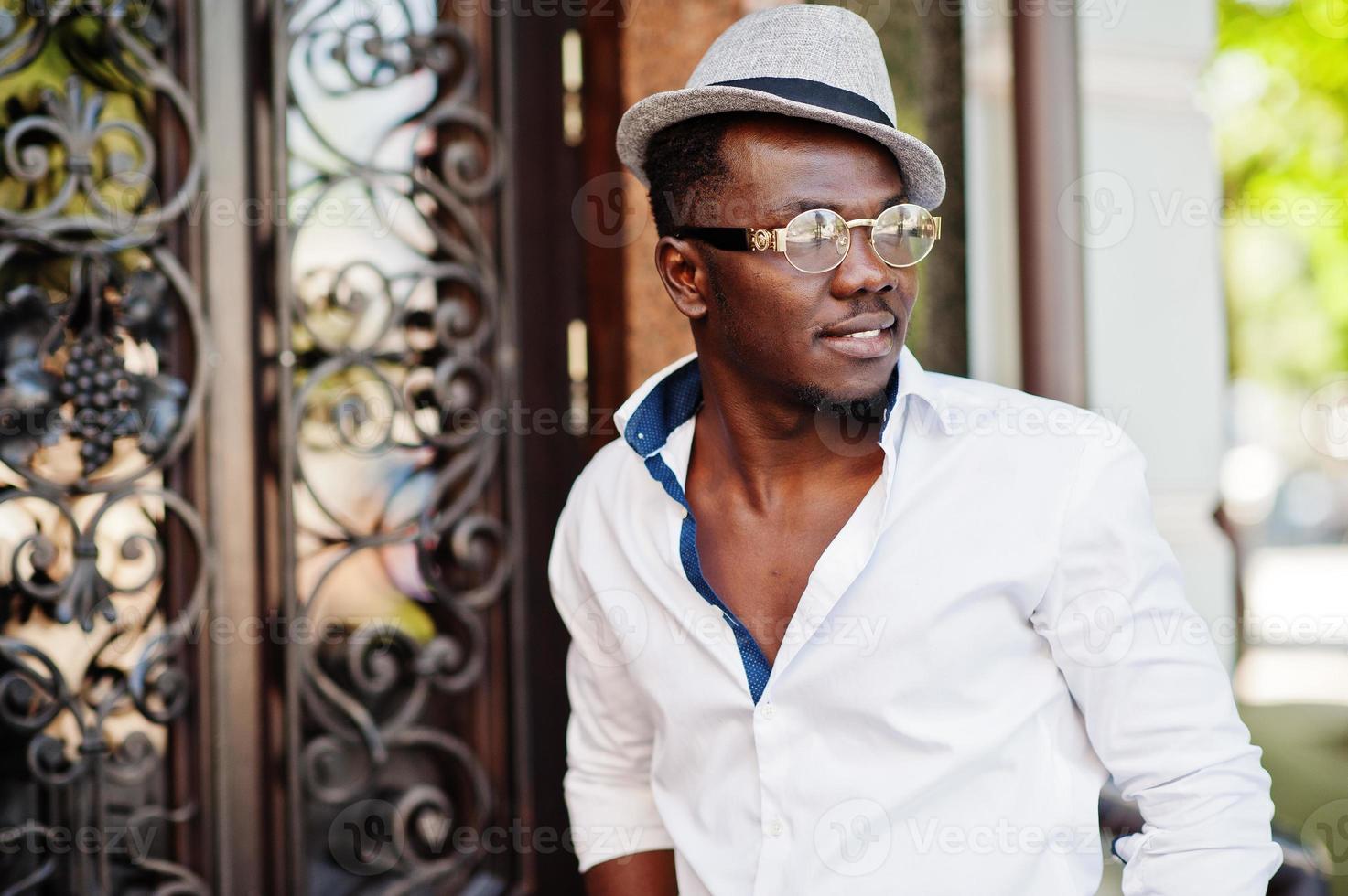 elegante uomo afroamericano in camicia bianca e pantaloni colorati con cappello e occhiali in posa all'aperto. ragazzo nero modello alla moda. foto