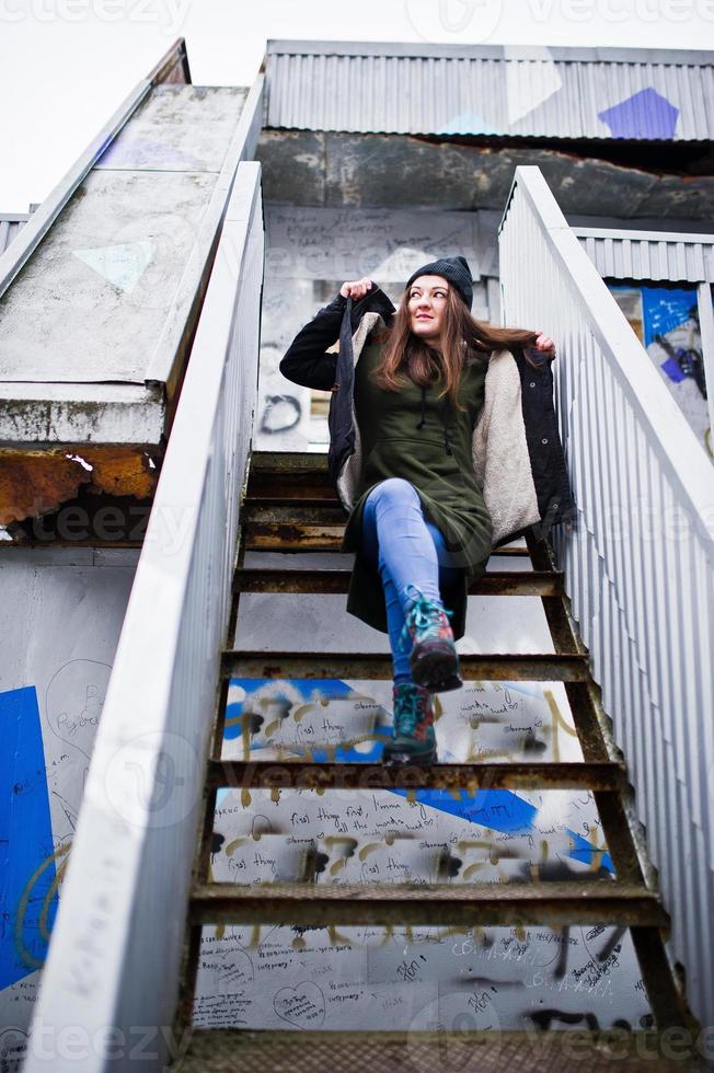 giovane ragazza indossa una lunga felpa verde, jeans e copricapo nero sul posto idustrial. foto