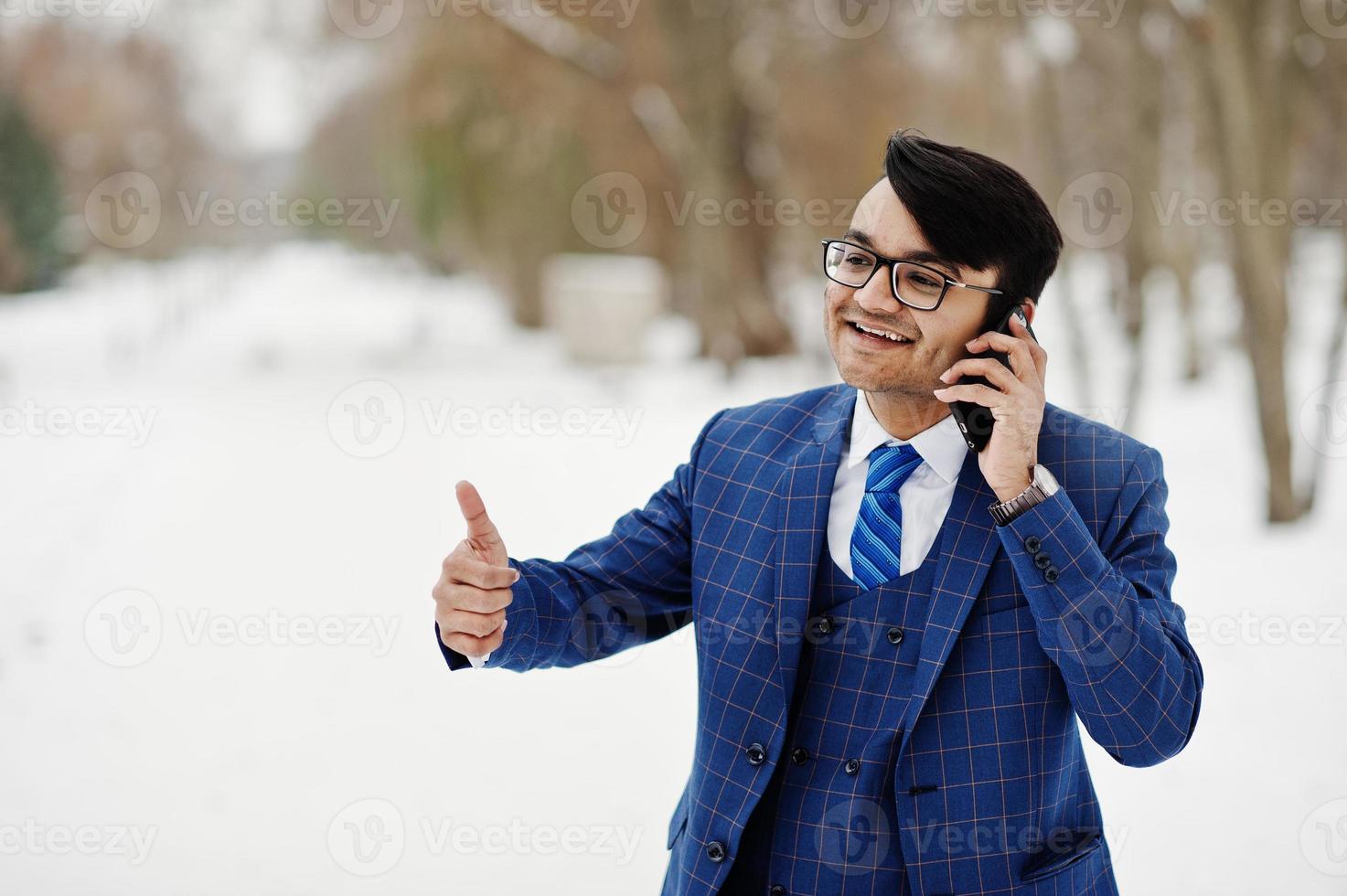 elegante uomo d'affari indiano in giacca e occhiali in posa al giorno d'inverno all'aperto, parlando sul telefono cellulare. foto