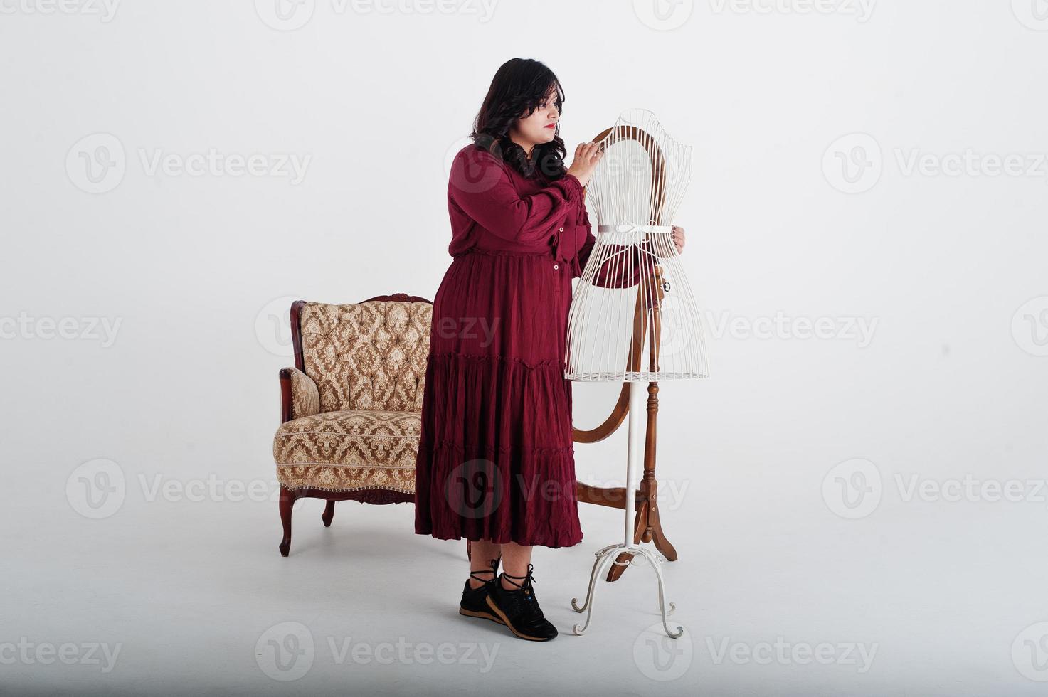 attraente donna del sud asiatico in abito rosso profondo dell'abito poste in studio su sfondo bianco contro specchio e sedia. foto