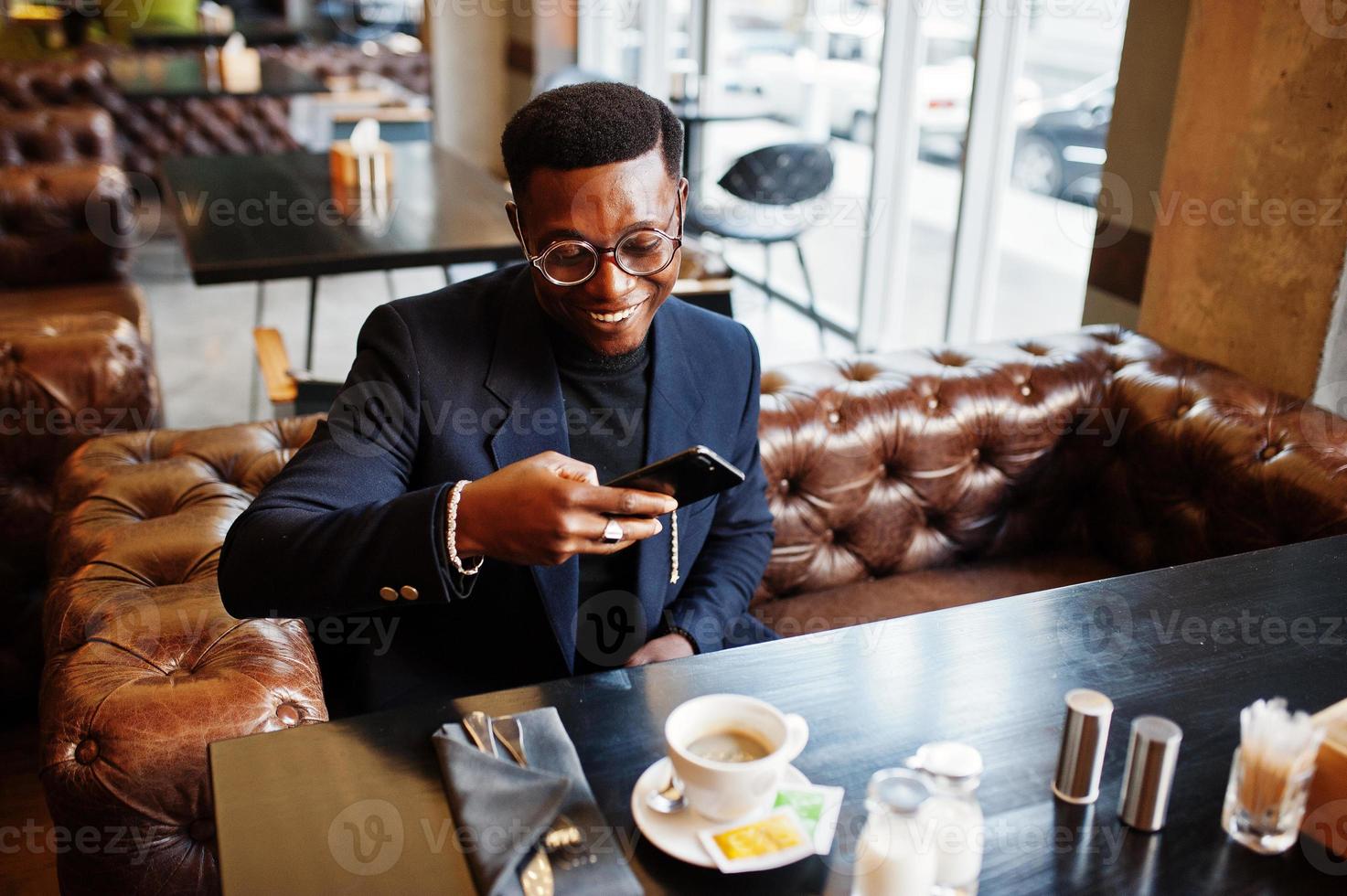 uomo afroamericano alla moda in giacca e occhiali seduto al bar e prendere un caffè al telefono. foto