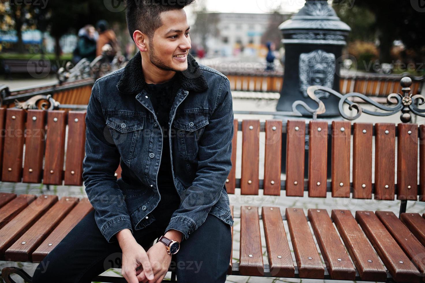 uomo indiano bello e alla moda in giacca di jeans neri in posa all'aperto, seduto su una panchina. foto