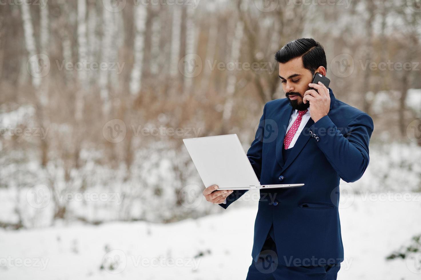 elegante uomo d'affari con barba indiana in giacca e cravatta posato in una giornata invernale all'aperto con un laptop a portata di mano, parlando sul telefono cellulare. foto