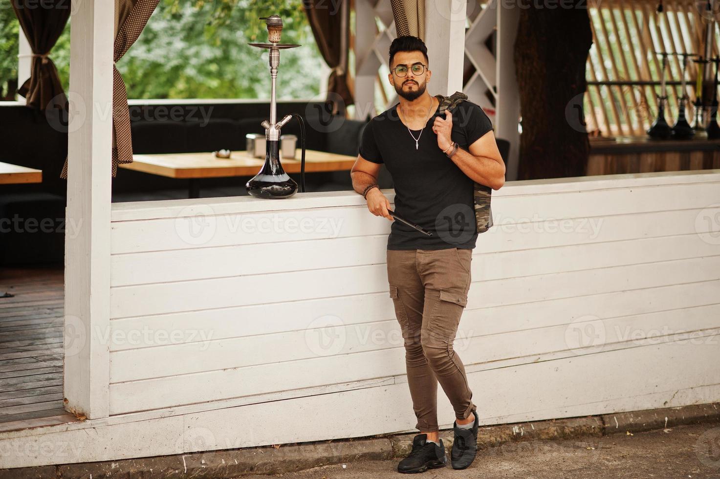 elegante barba uomo arabo in occhiali e t-shirt nera che fuma narghilè all'aperto. modello arabo che riposa. foto