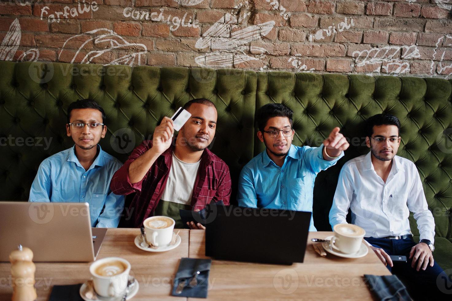 un gruppo di quattro uomini del sud asiatico poste alla riunione di lavoro nella caffetteria. gli indiani lavorano insieme con i laptop usando vari gadget, conversando. tenere la carta di credito per pagare. foto