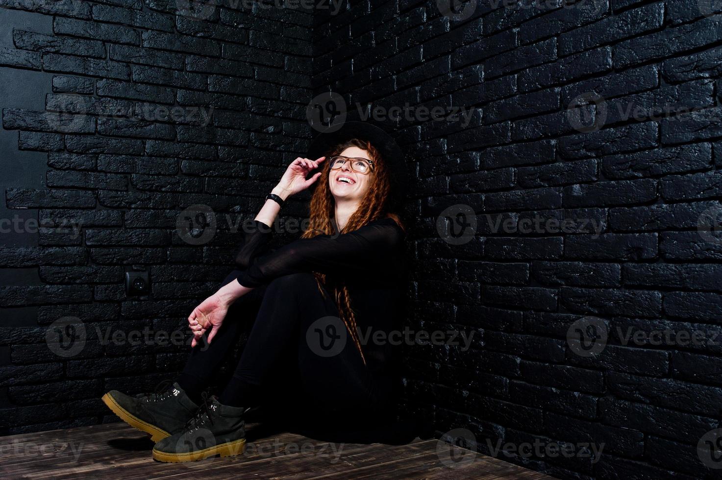 tiro in studio di una ragazza in nero con dreadlocks, occhiali e cappello su sfondo di mattoni. foto