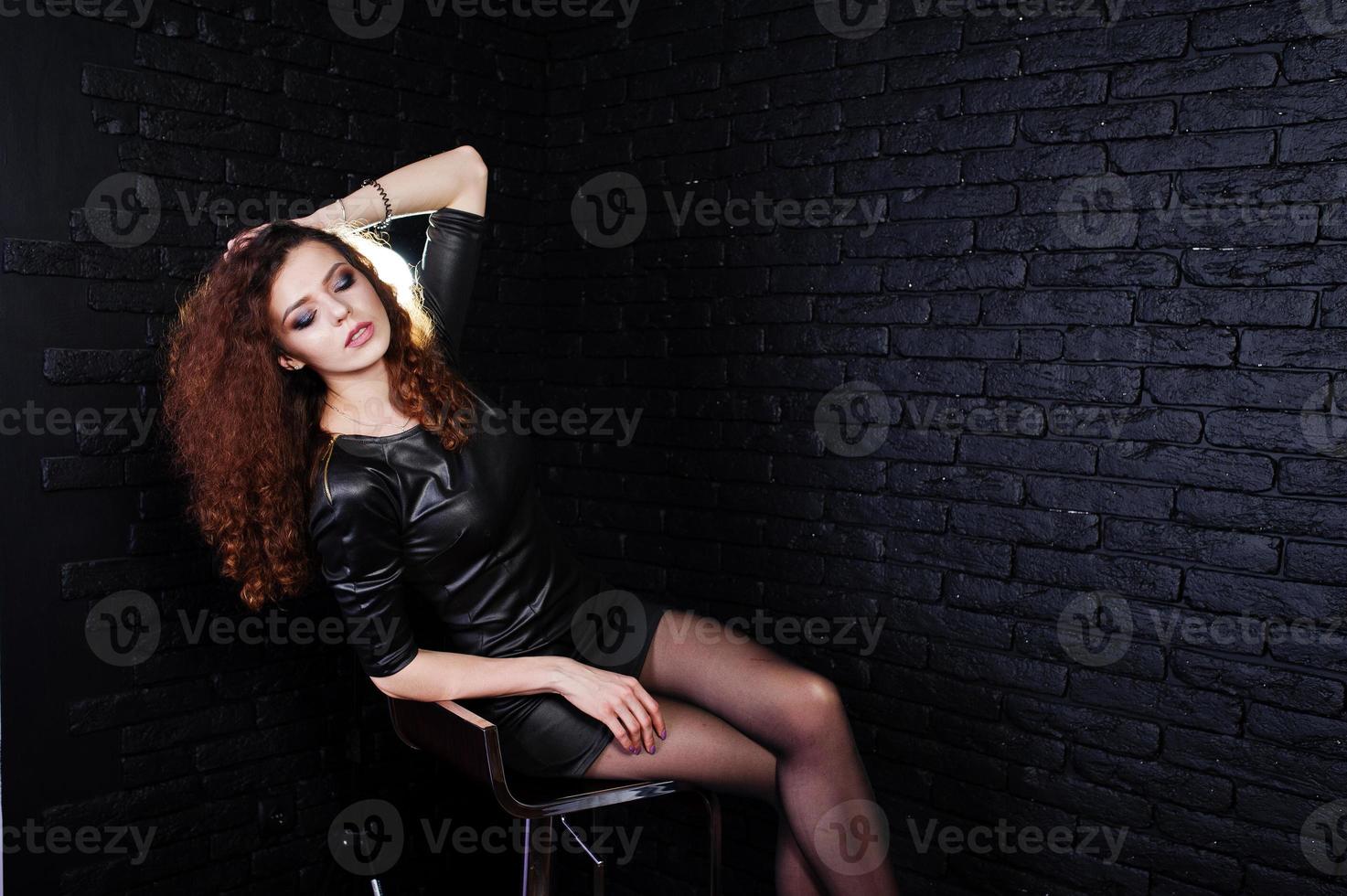 bruna dai capelli ricci gambe lunghe ragazza in abito di pelle nera poste in studio su una sedia contro un muro di mattoni scuri. foto