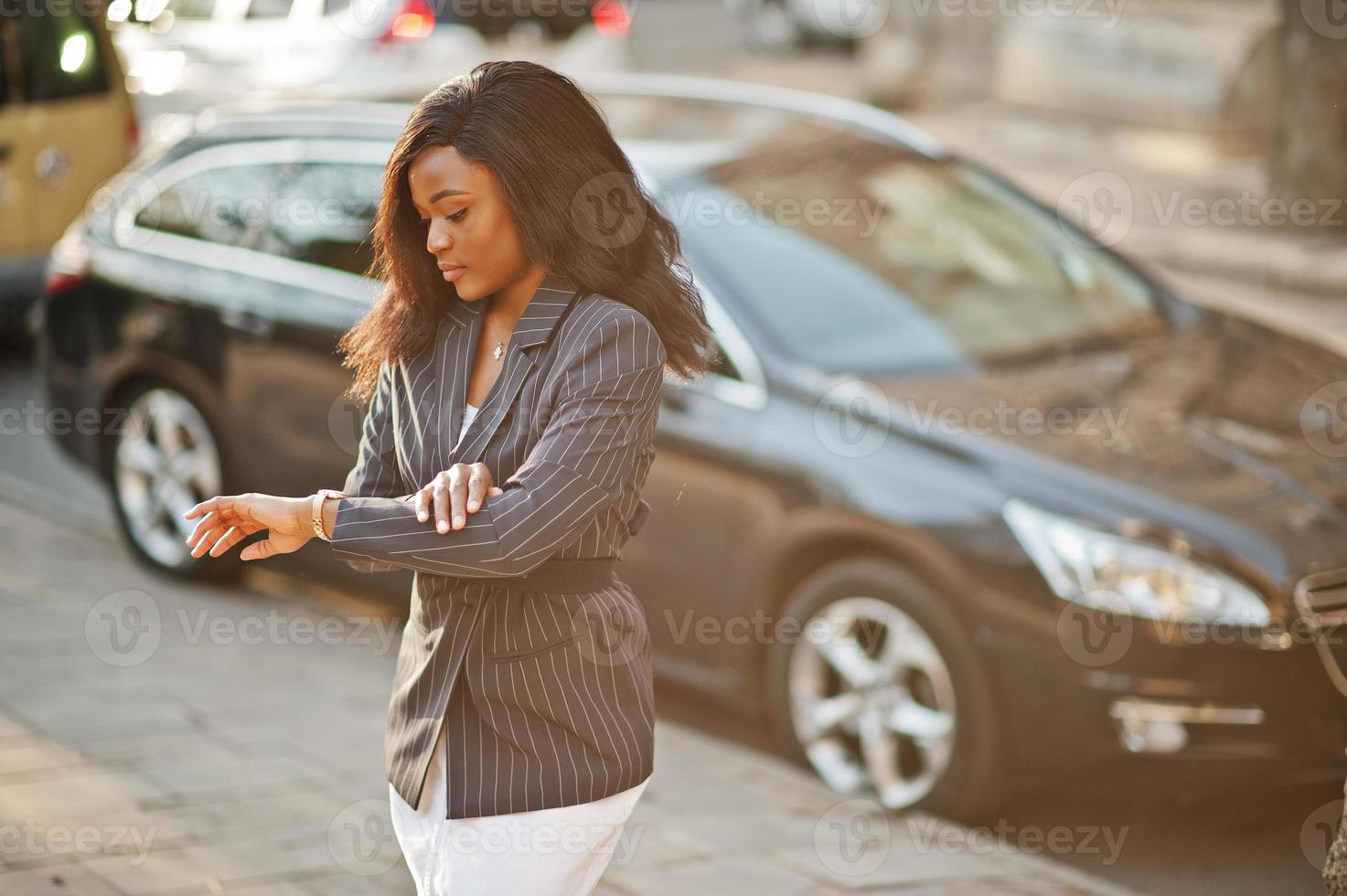 successo elegante donna afroamericana in giacca contro auto nera guardando i suoi orologi. foto