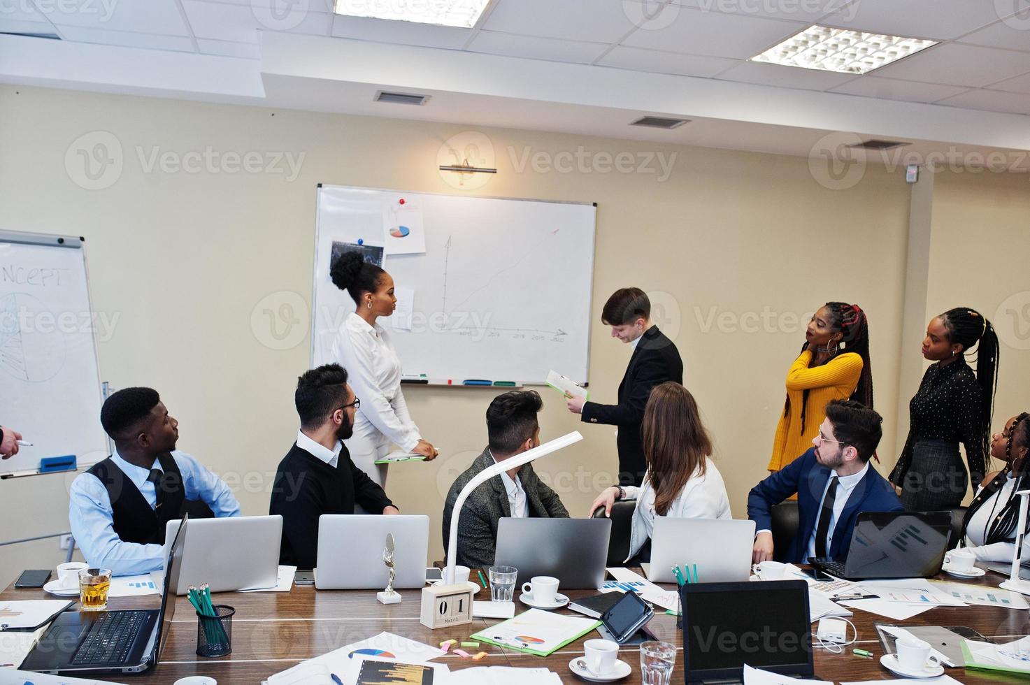 allenatore di affari di razza mista che presenta un rapporto in piedi vicino alla lavagna che indica le statistiche di vendita mostrate sul diagramma e sul grafico insegnano a diversi membri dell'azienda riuniti nella sala conferenze. foto