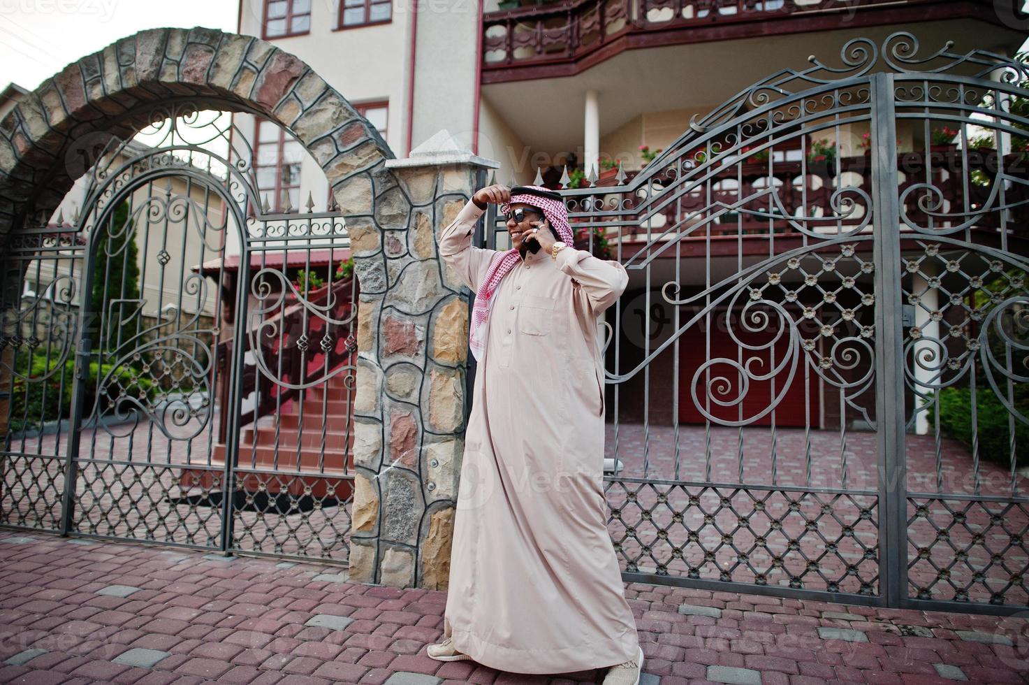 ricco uomo d'affari arabo mediorientale con occhiali da sole in posa per strada contro il palazzo, parlando al telefono cellulare. foto
